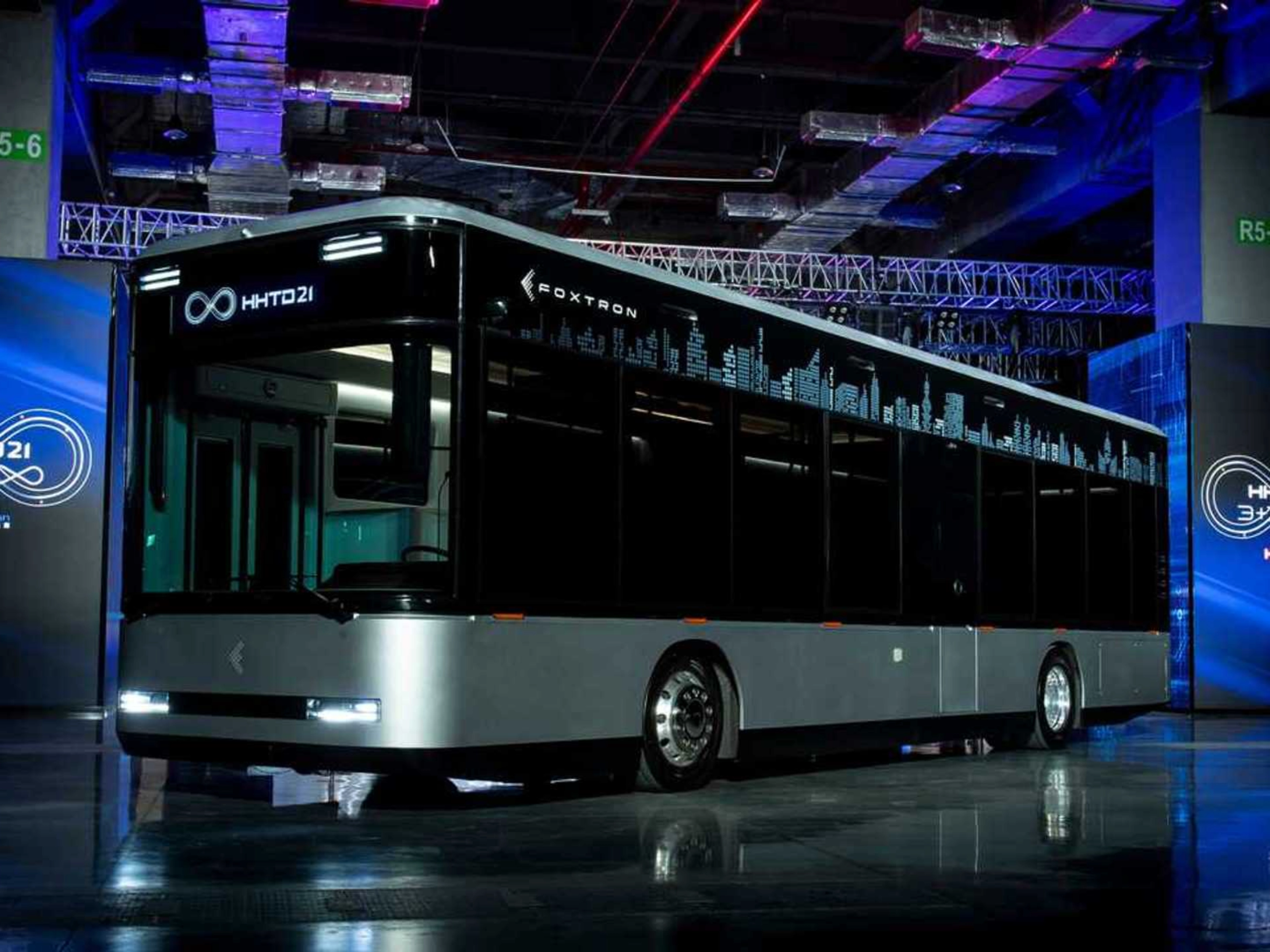 A modern electric city bus is showcased indoors with the logo "Foxtron" and digital skyline.