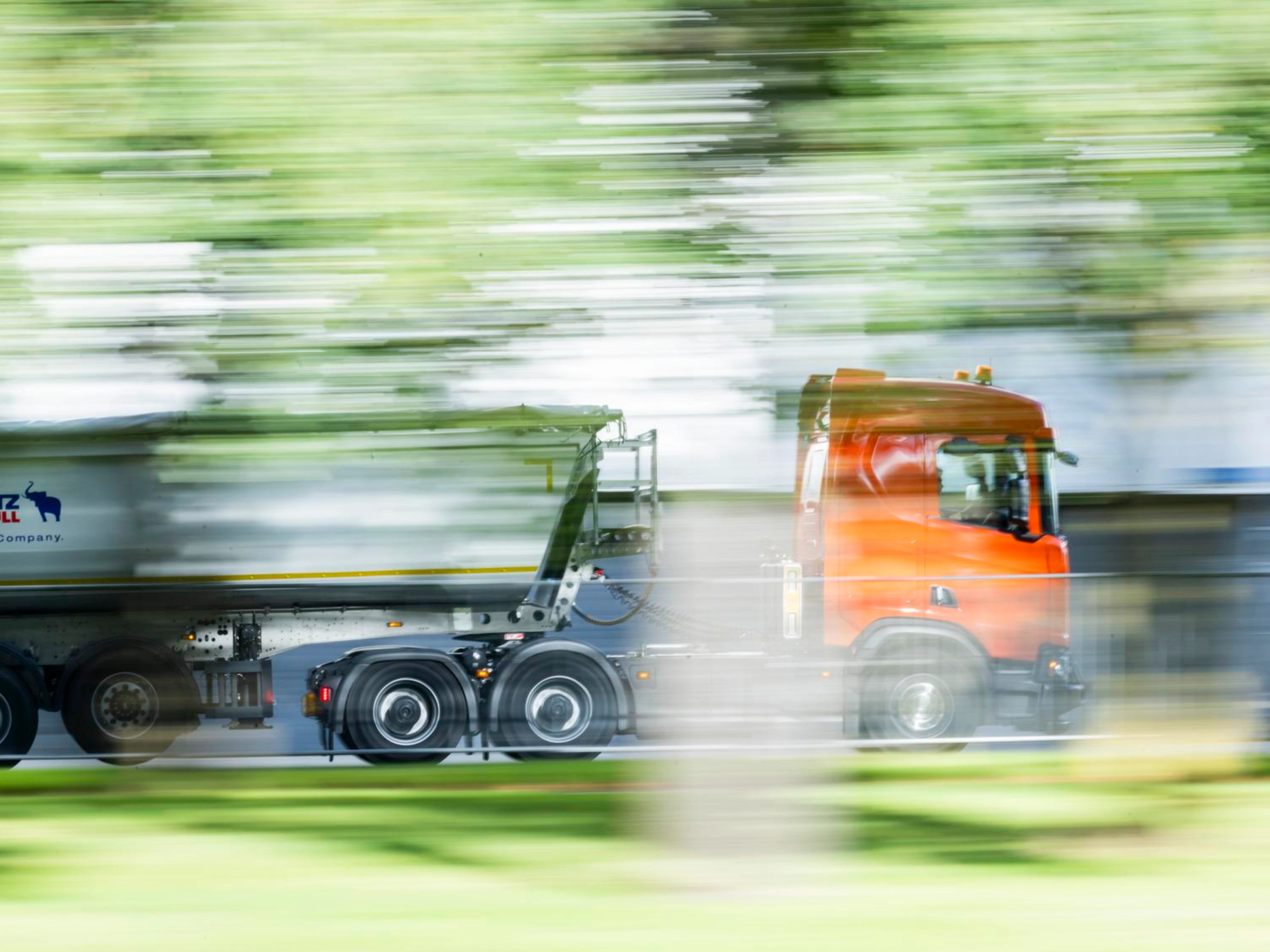 A truck with a trailer drives along a road surrounded by greenery.