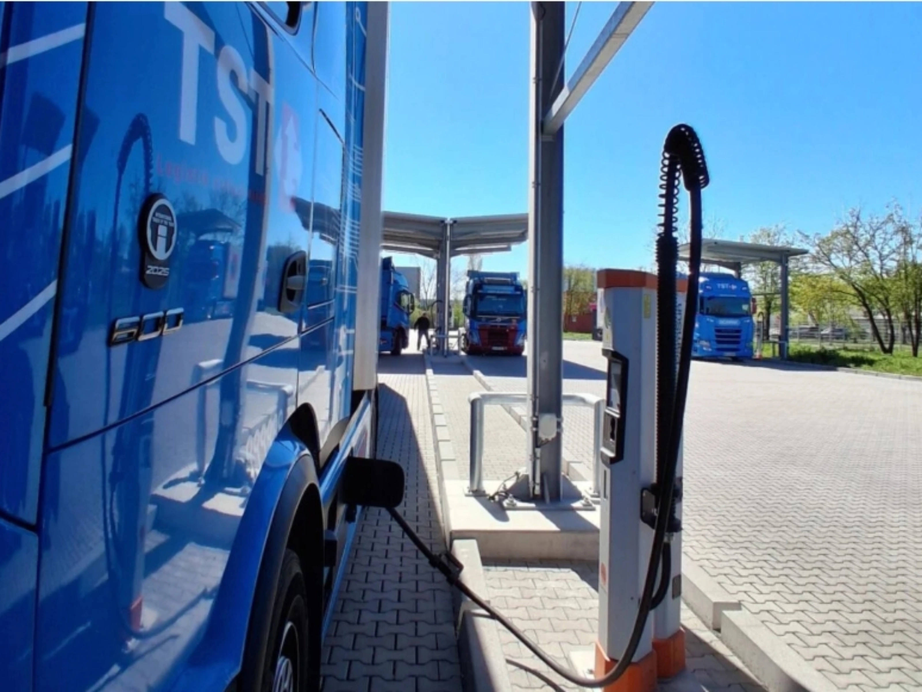 Blue electric truck connected to a charging station at a parking area.