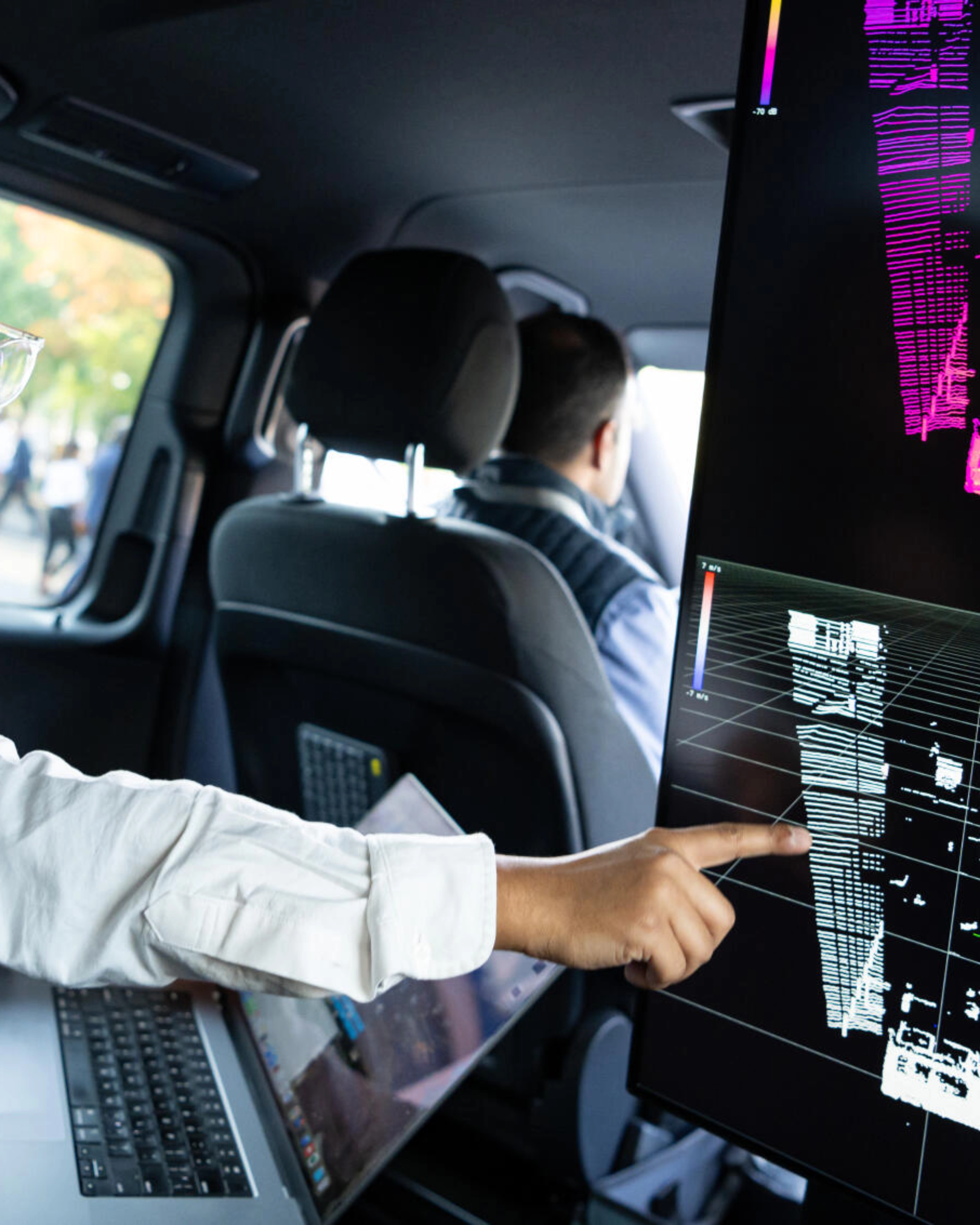 Two men in a car analyze 3D sensor data on a screen, likely testing autonomous driving tech.