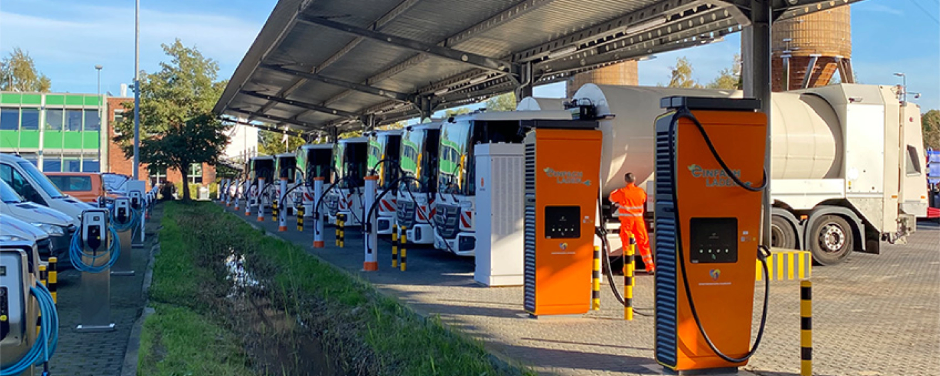Orange charging stations beneath a solar canopy at a parking area with parked trucks.