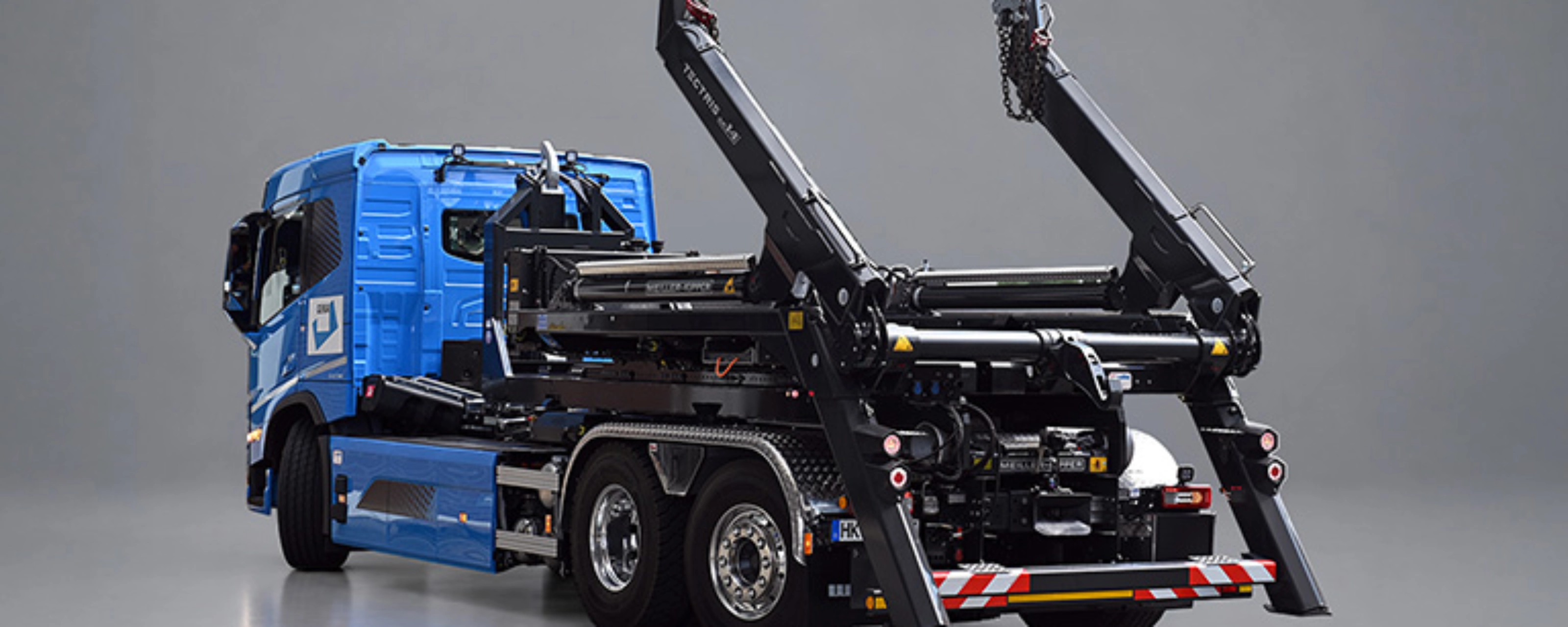 Blauer Lkw mit schwarzem Meiller-Hakenliftsystem und angehobenen Hubarmen in schräger Rückansicht.