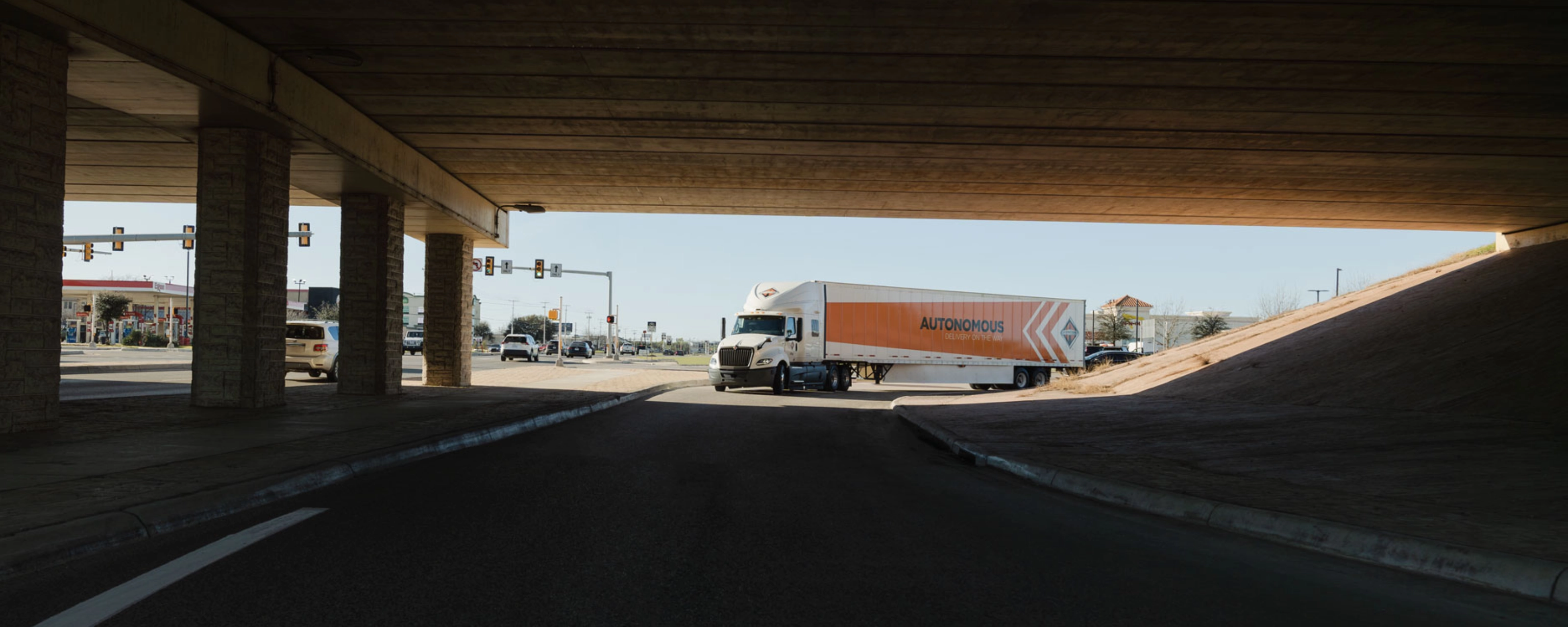 A self-driving truck with an "Autonomous" trailer drives through an intersection under a bridge.