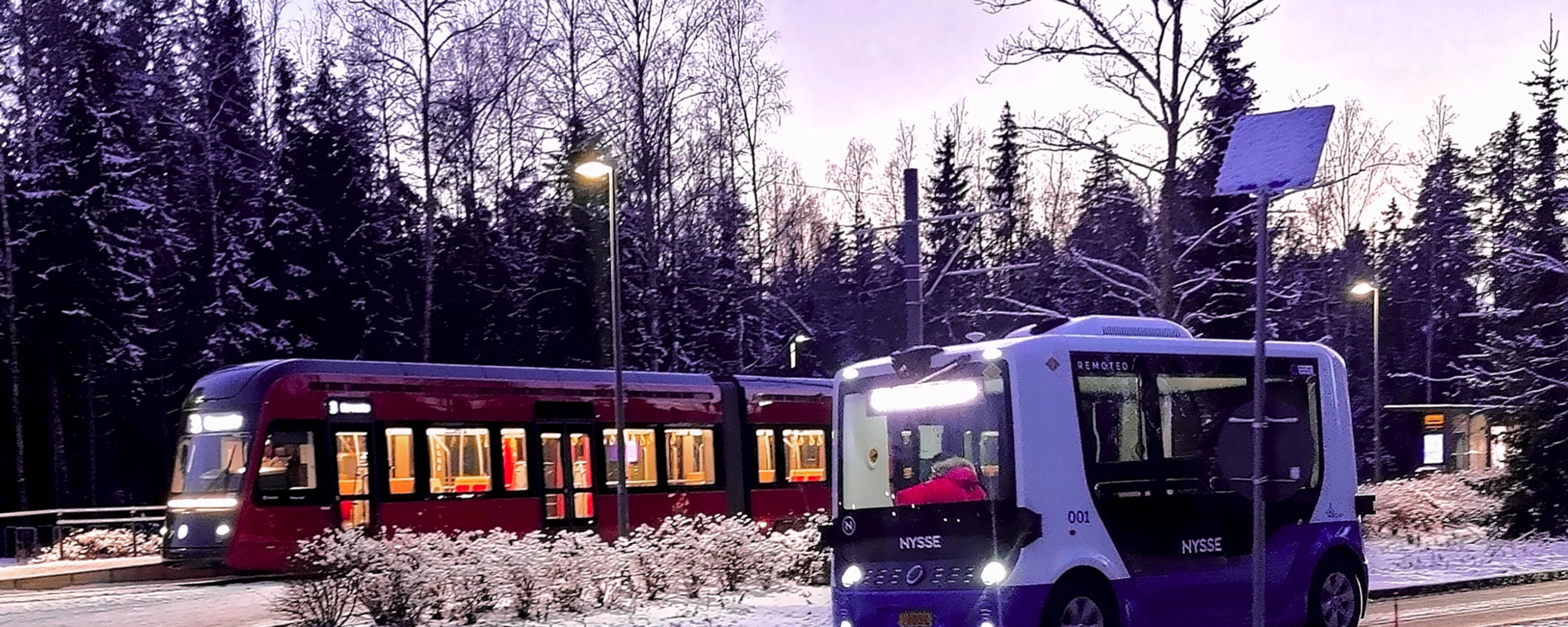 Autonomous shuttle bus driving beside a tram on a snowy street.