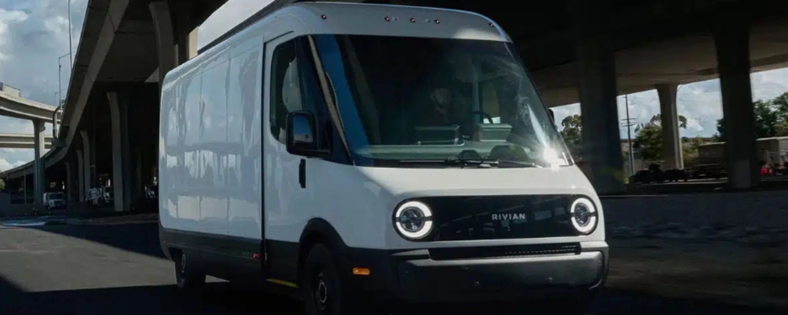White Rivian electric delivery van driving on a city street under a highway bridge.