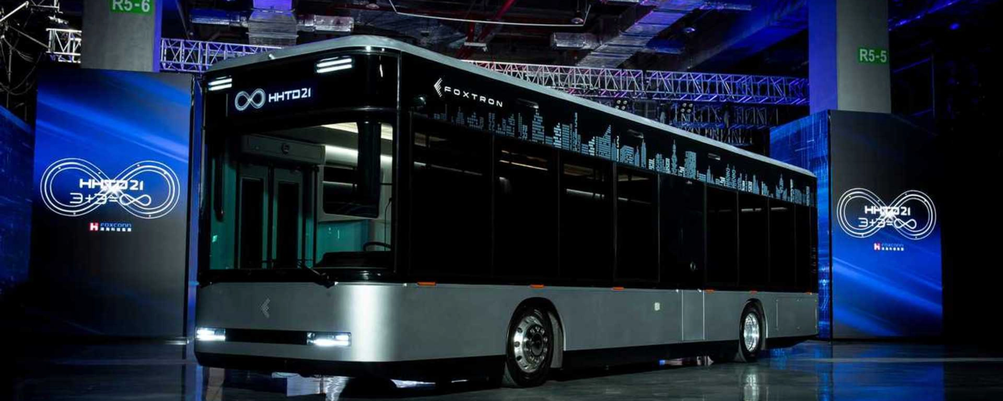 A modern electric city bus is showcased indoors with the logo "Foxtron" and digital skyline.
