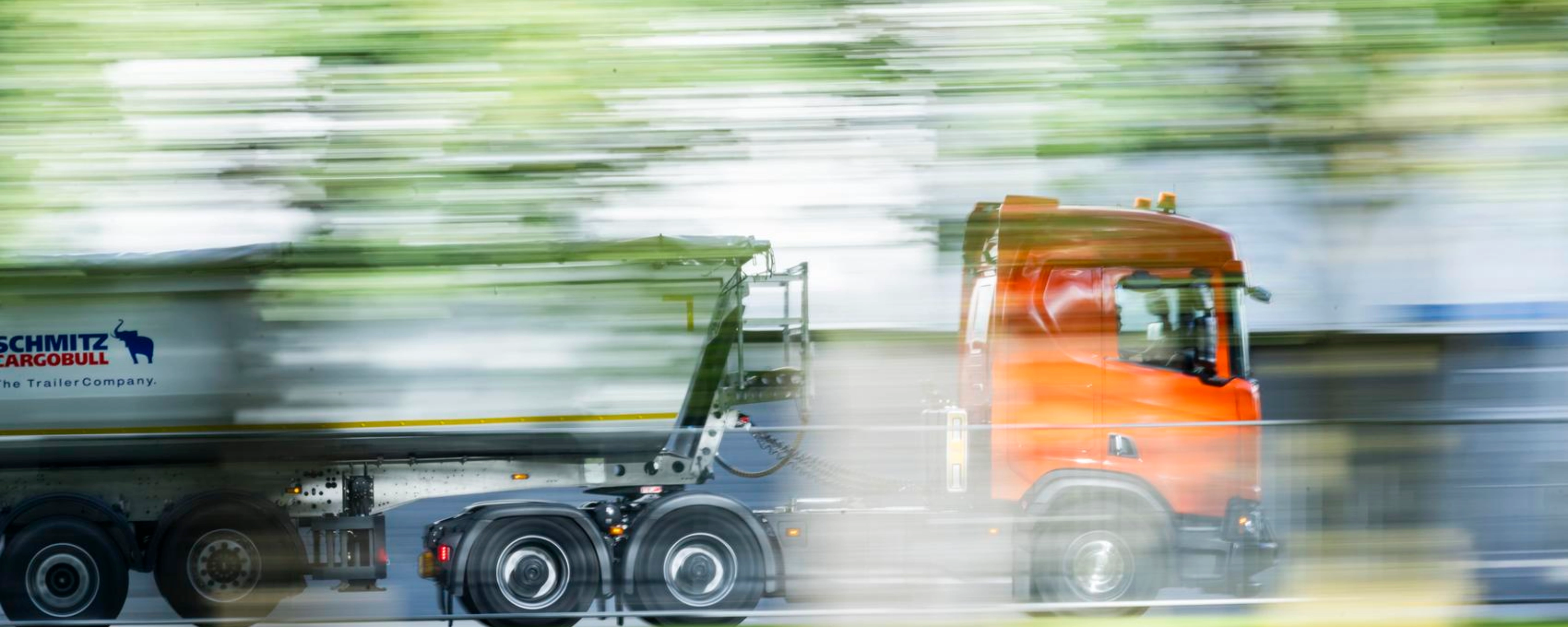 A truck with a trailer drives along a road surrounded by greenery.