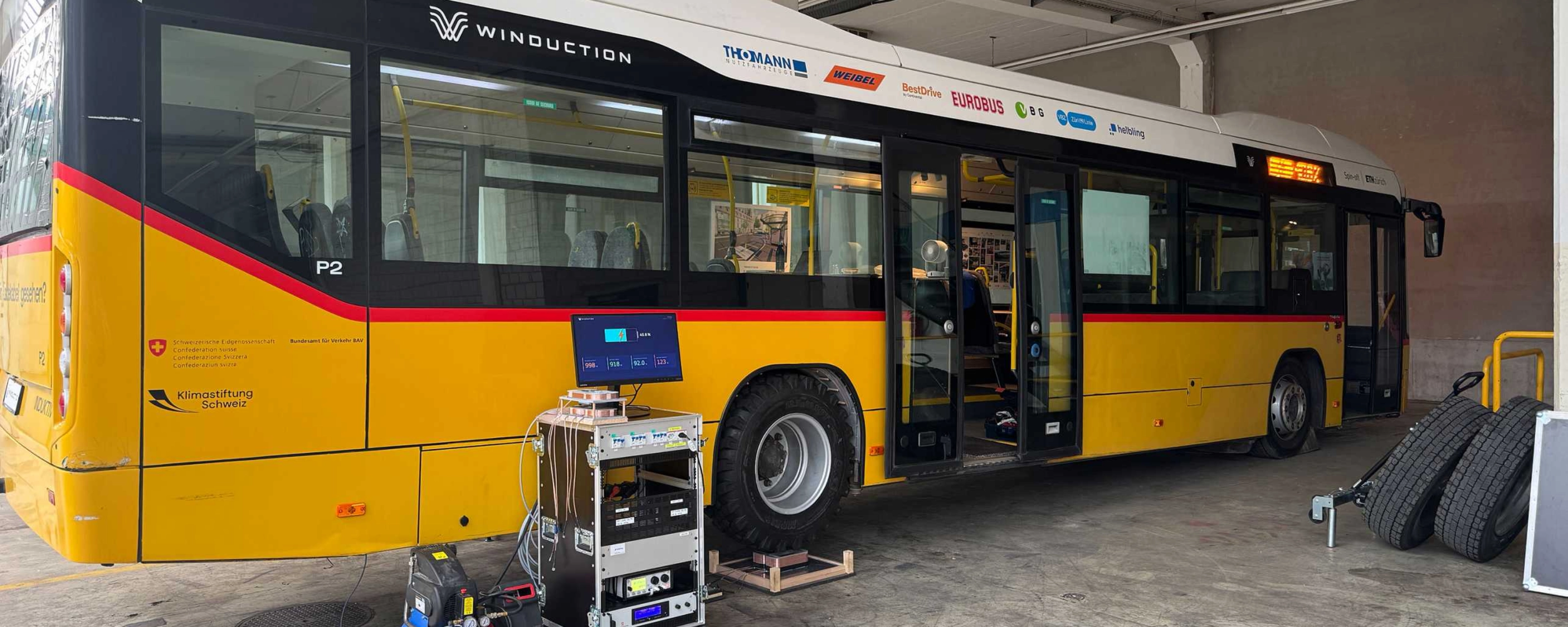 Yellow electric bus in a workshop with open doors and technical equipment in front of it.