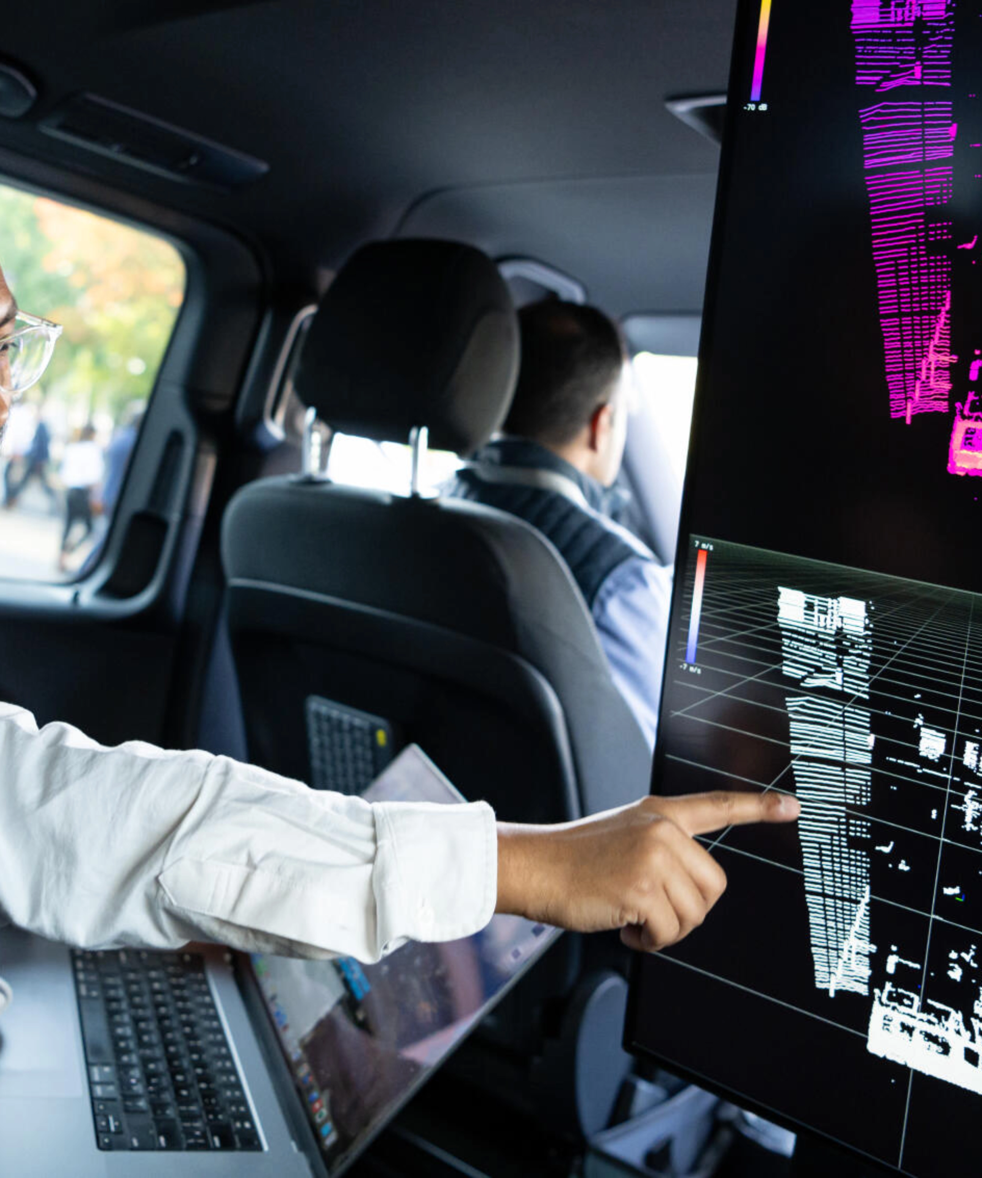 Two men in a car analyze 3D sensor data on a large screen for autonomous driving.