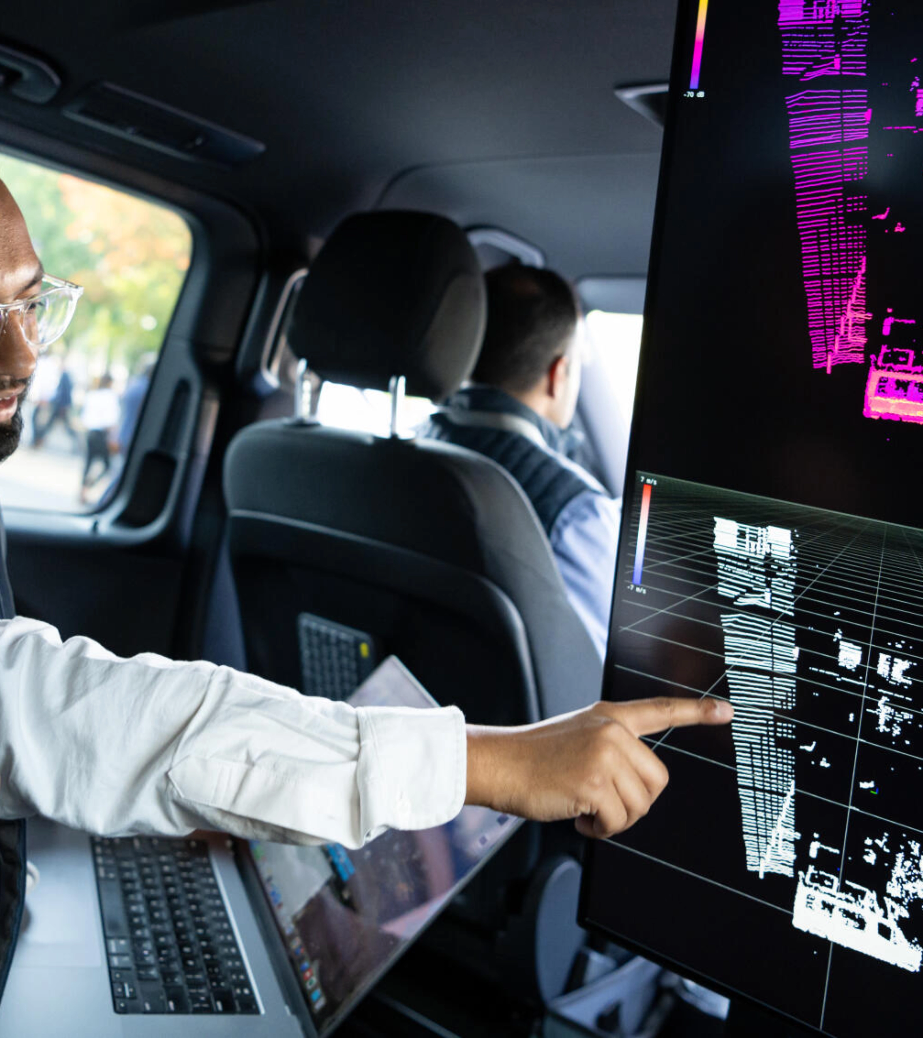 Two men in a car analyze 3D sensor data on a screen, likely testing autonomous driving tech.
