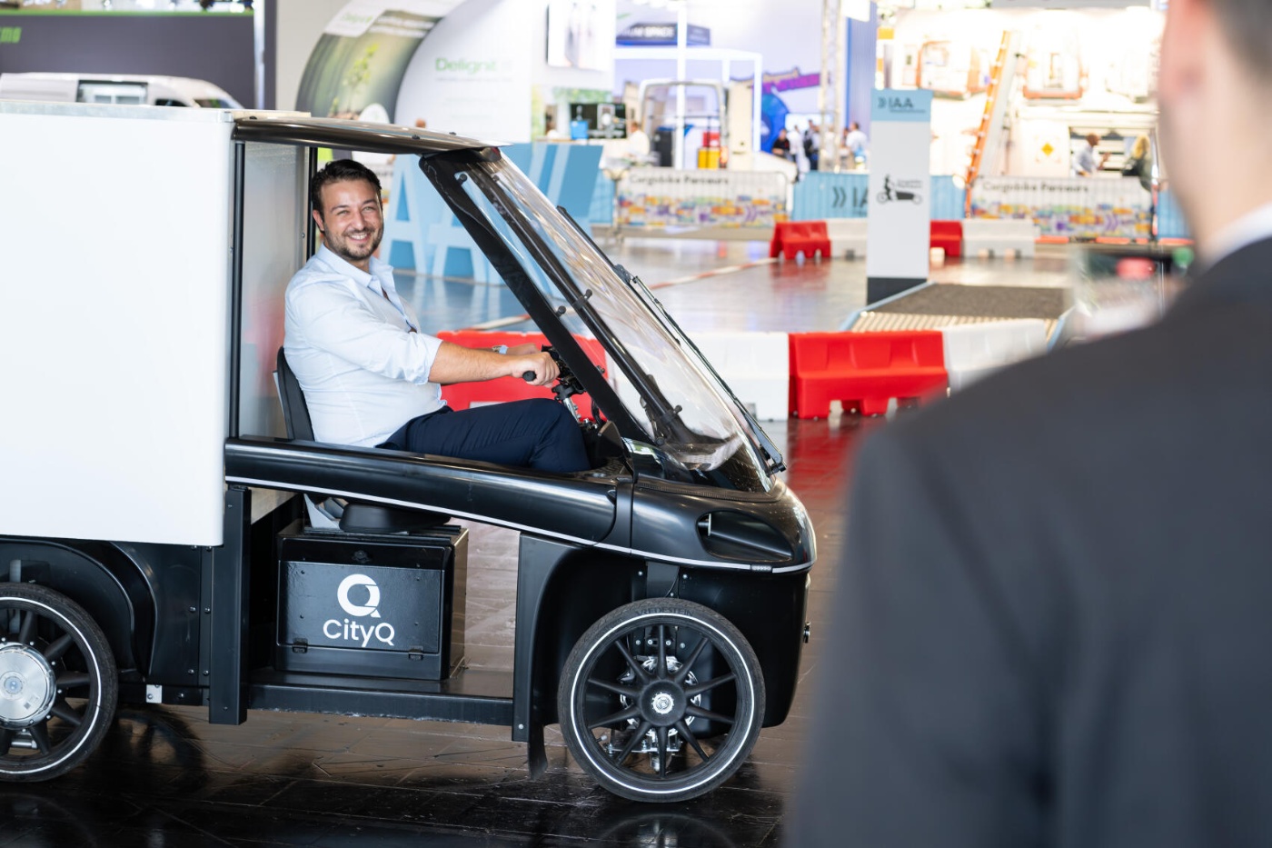 Person sitzt in einem kleinen elektrischen Lieferfahrzeug, während eine weitere Person daneben steht.