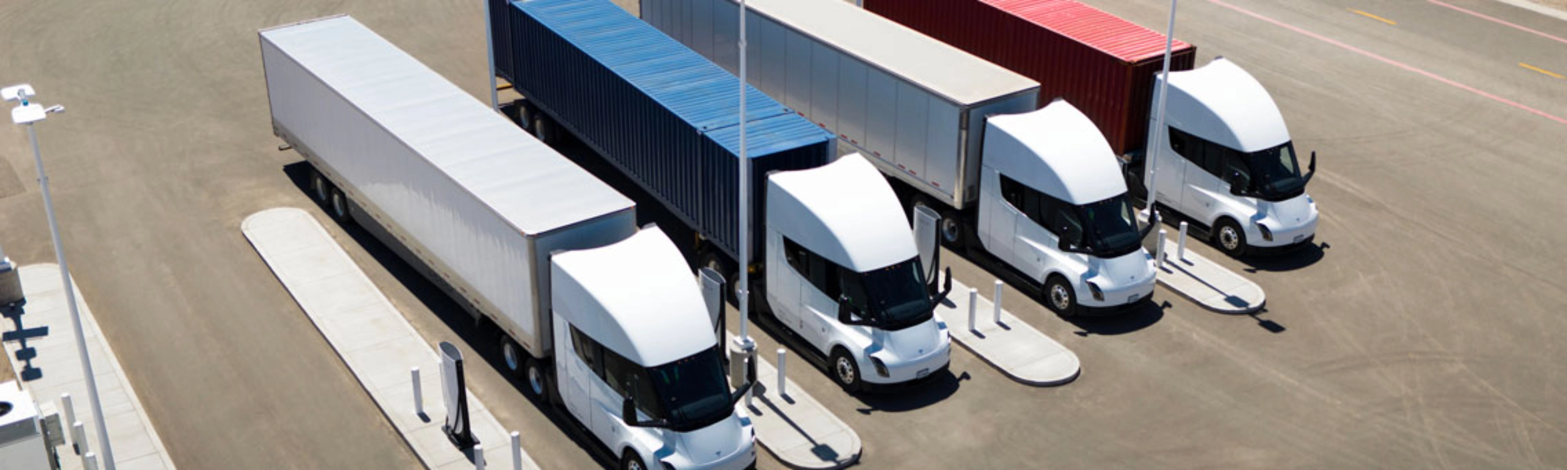 Several white electric semi-trucks are charging at a large station with colored containers.