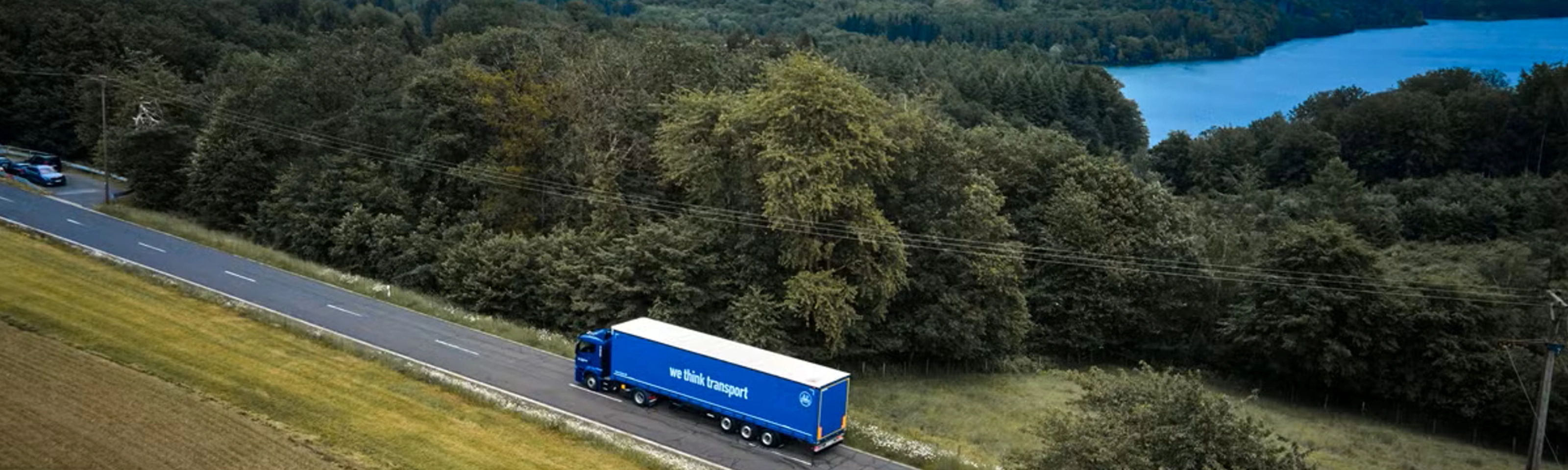 Blue BPW truck driving on a rural road beside a forest and lake.
