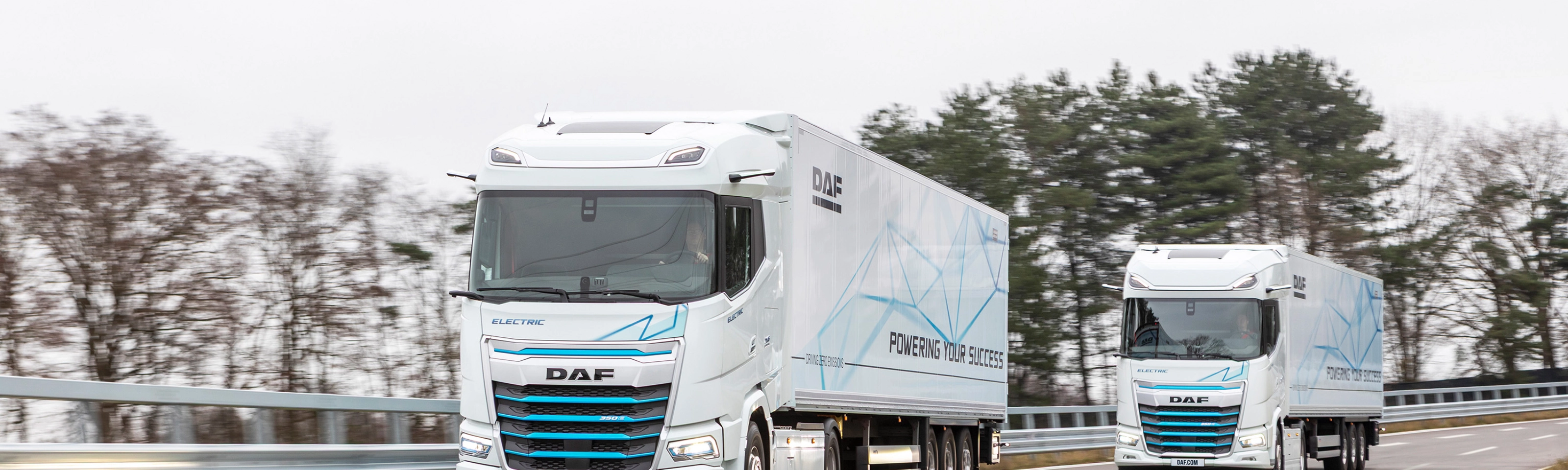 Two electric DAF trucks driving on a curved road with trees in the background.