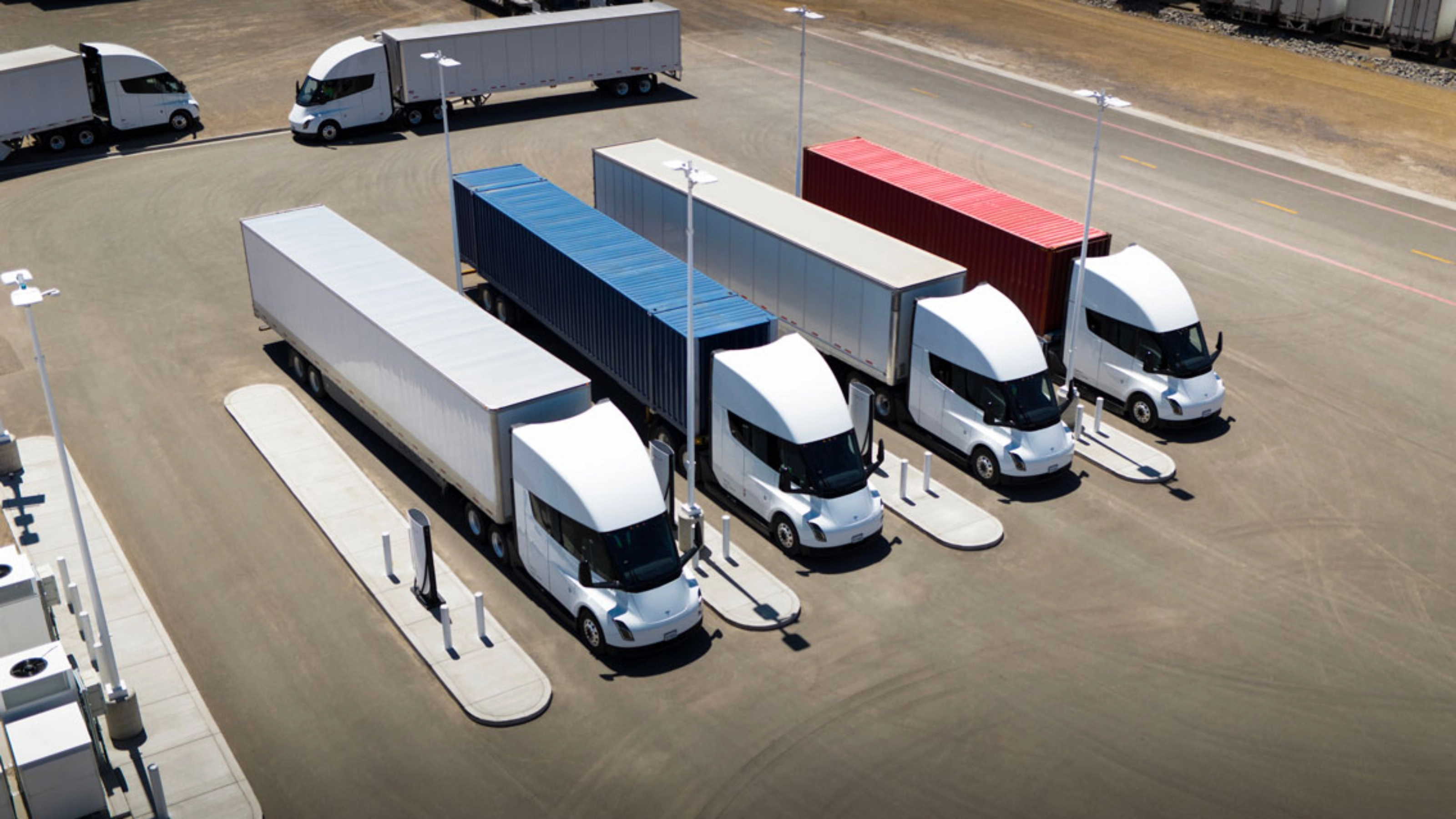 Several white electric semi-trucks are charging at a large station with colored containers.
