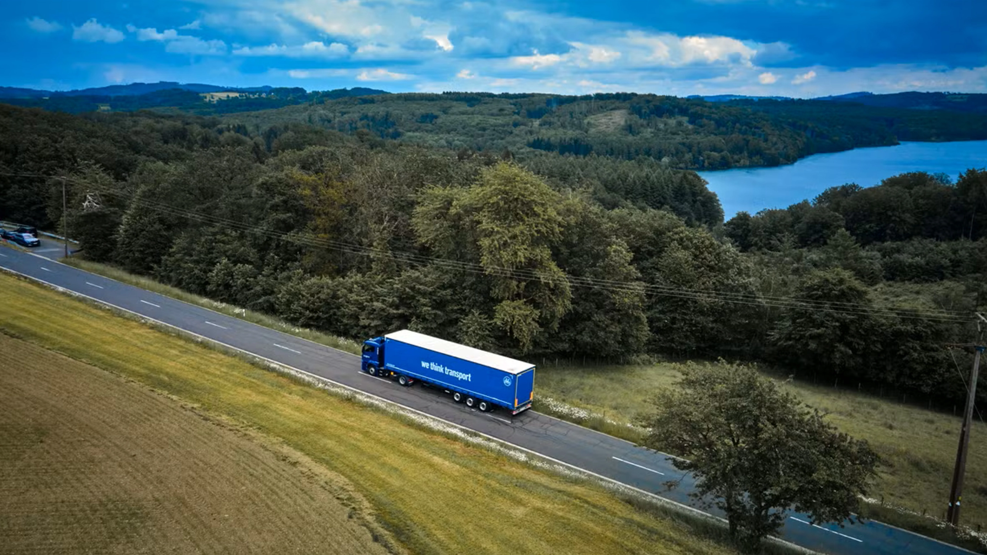 Blue BPW truck driving on a rural road beside a forest and lake.