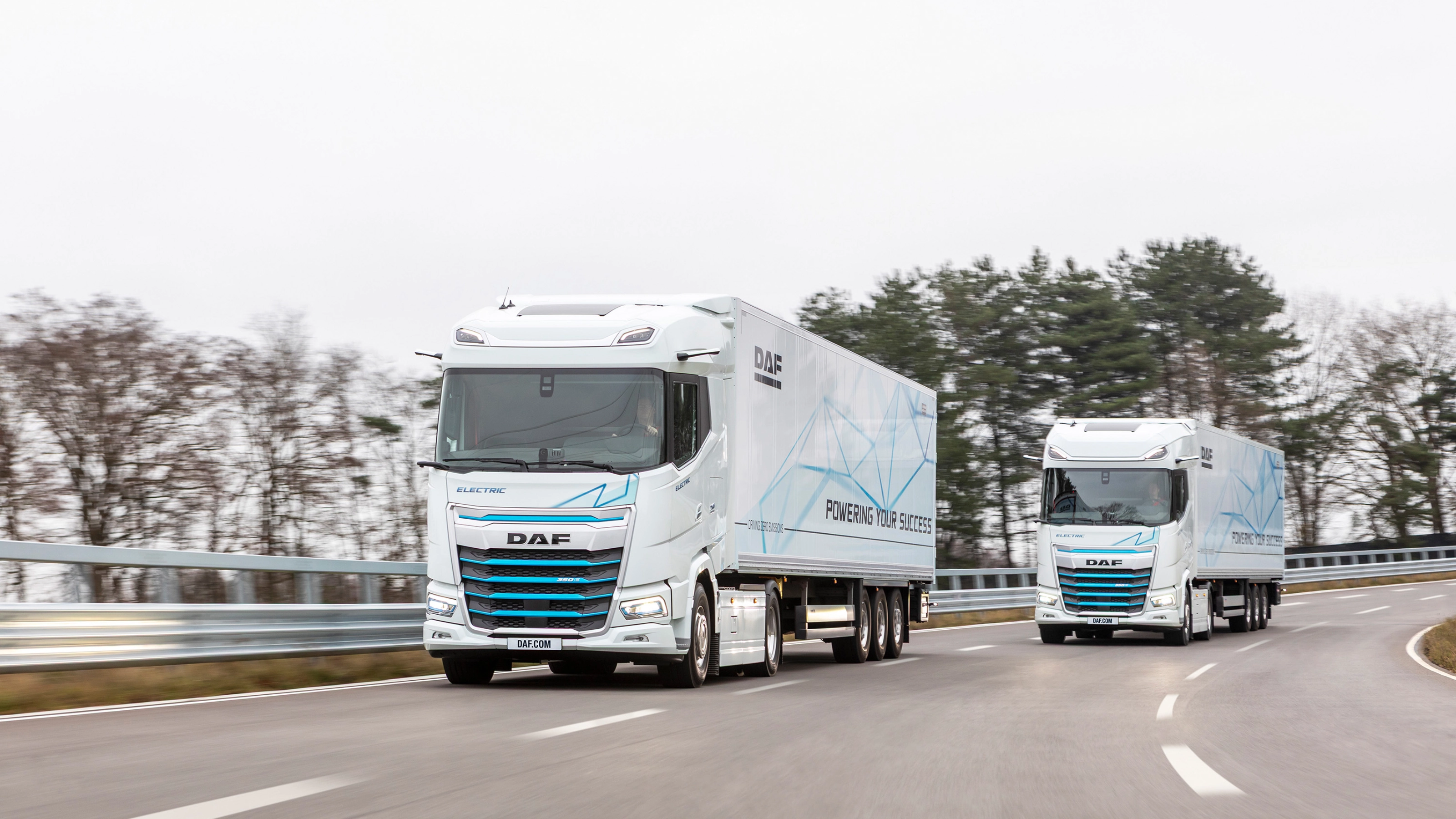 Two electric DAF trucks driving on a curved road with trees in the background.