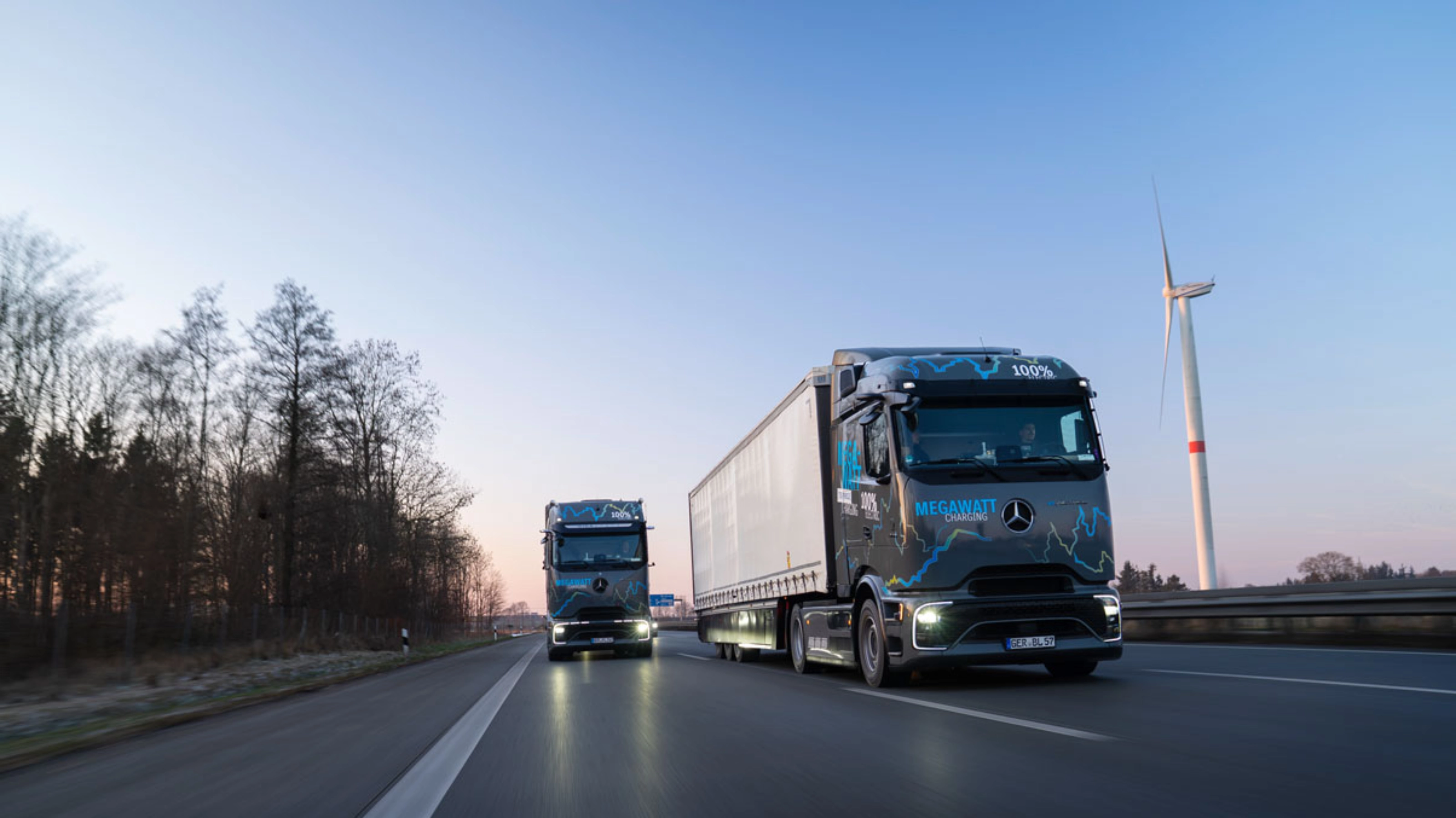 Zwei elektrische Mercedes-Benz-Lkw mit „Megawatt Charging“-Aufschrift fahren bei Dämmerung auf der Autobahn.