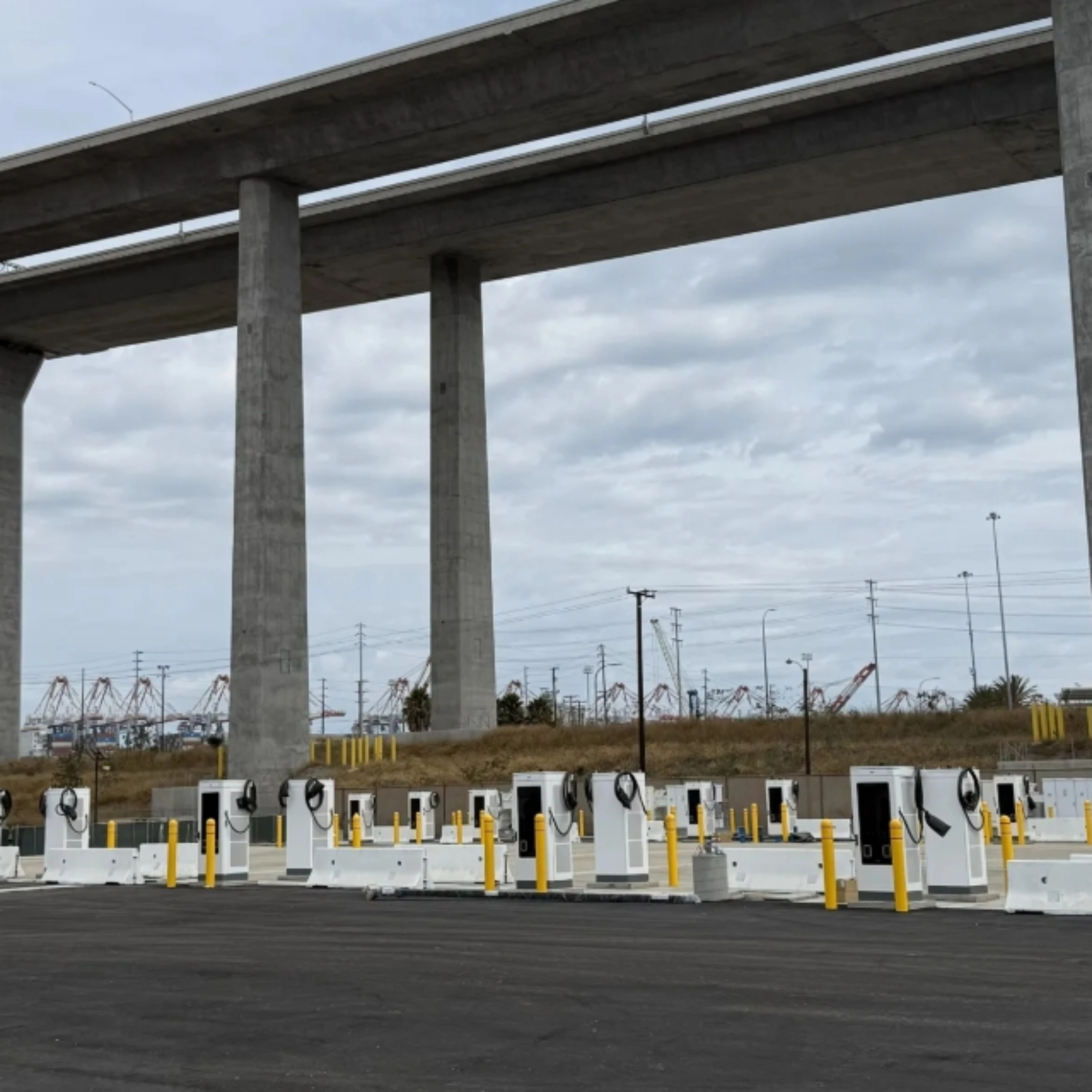 Mehrere E-Ladestationen auf einem Parkplatz unter einer Autobahnbrücke.