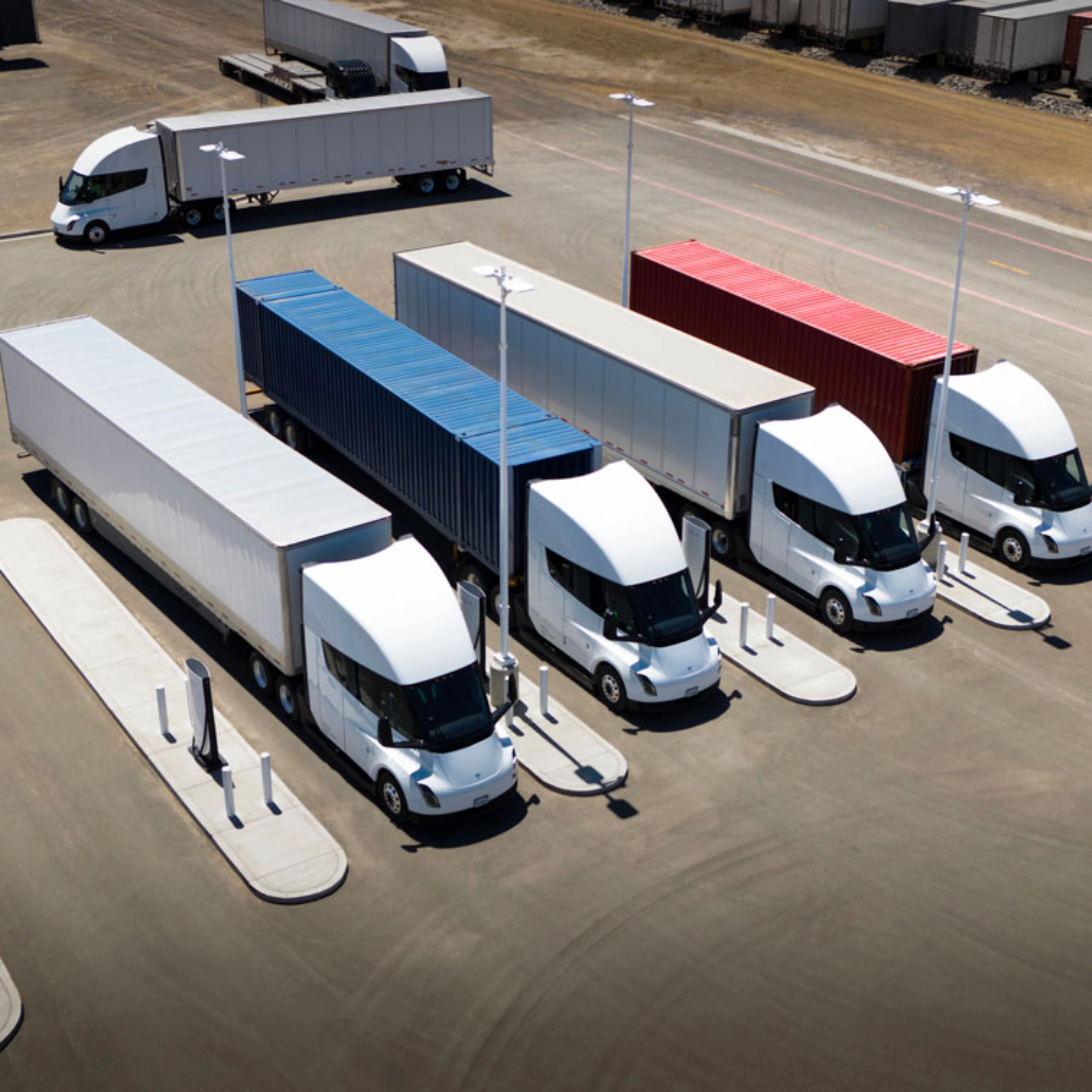 Several white electric semi-trucks are charging at a large station with colored containers.