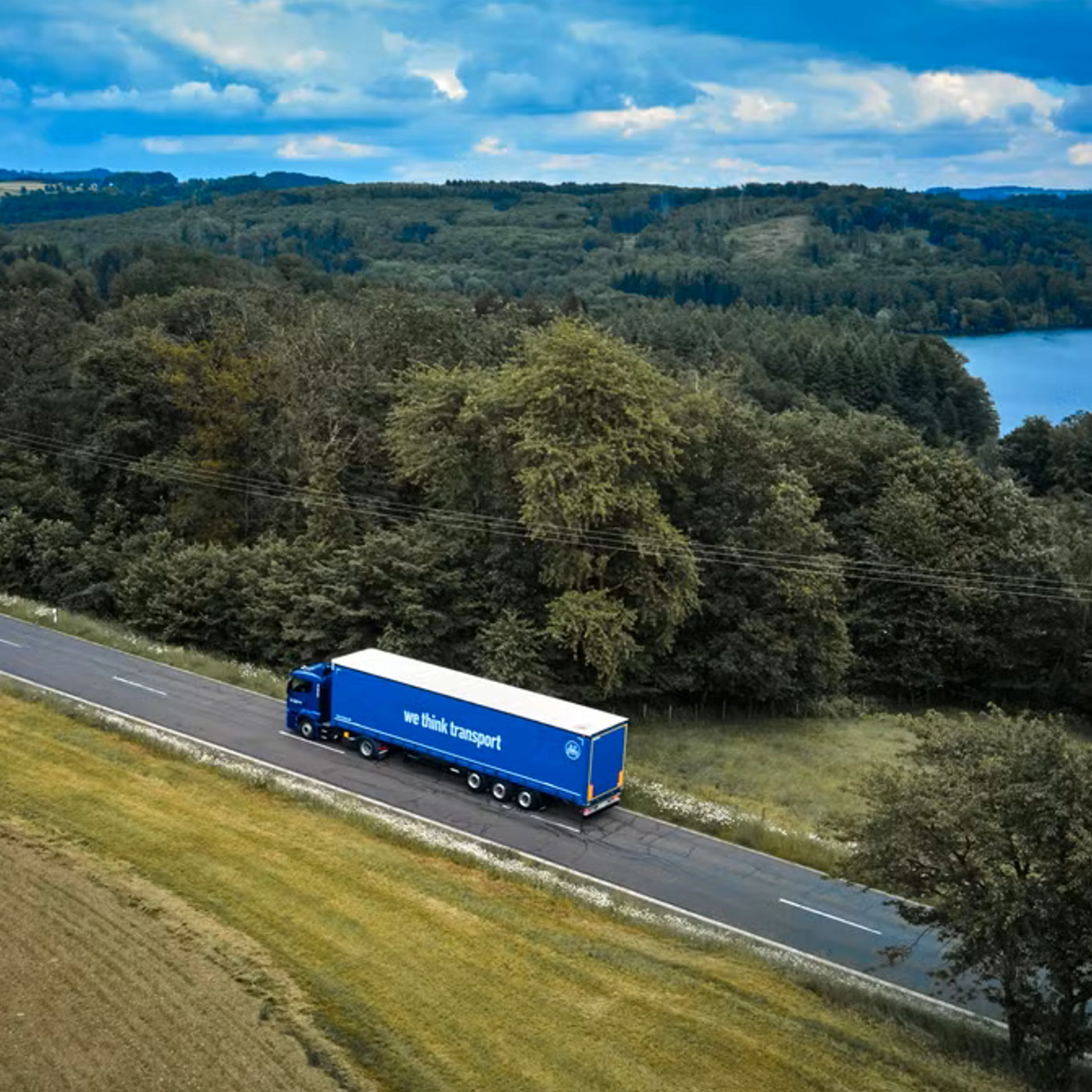 Blue BPW truck driving on a rural road beside a forest and lake.