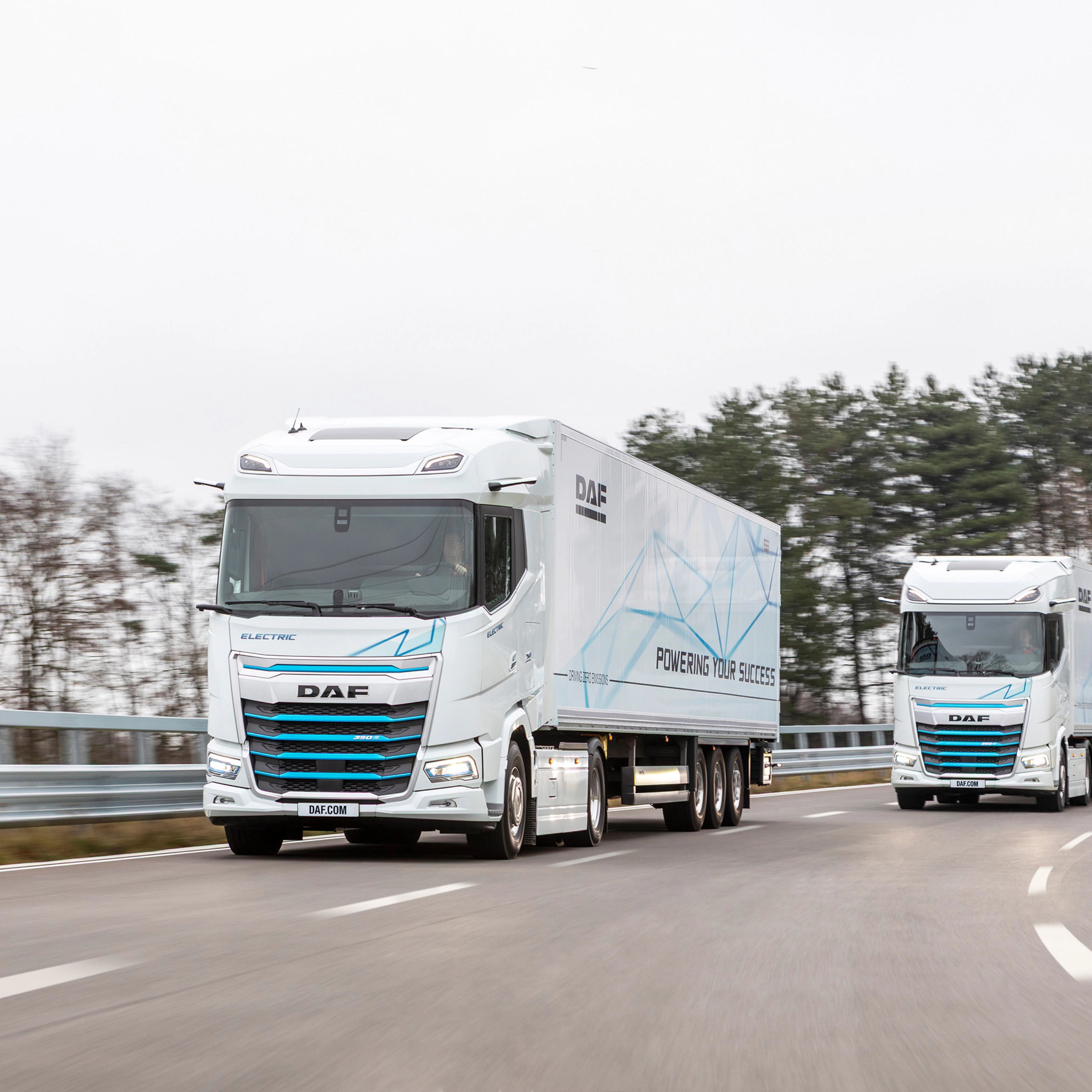 Two electric DAF trucks driving on a curved road with trees in the background.