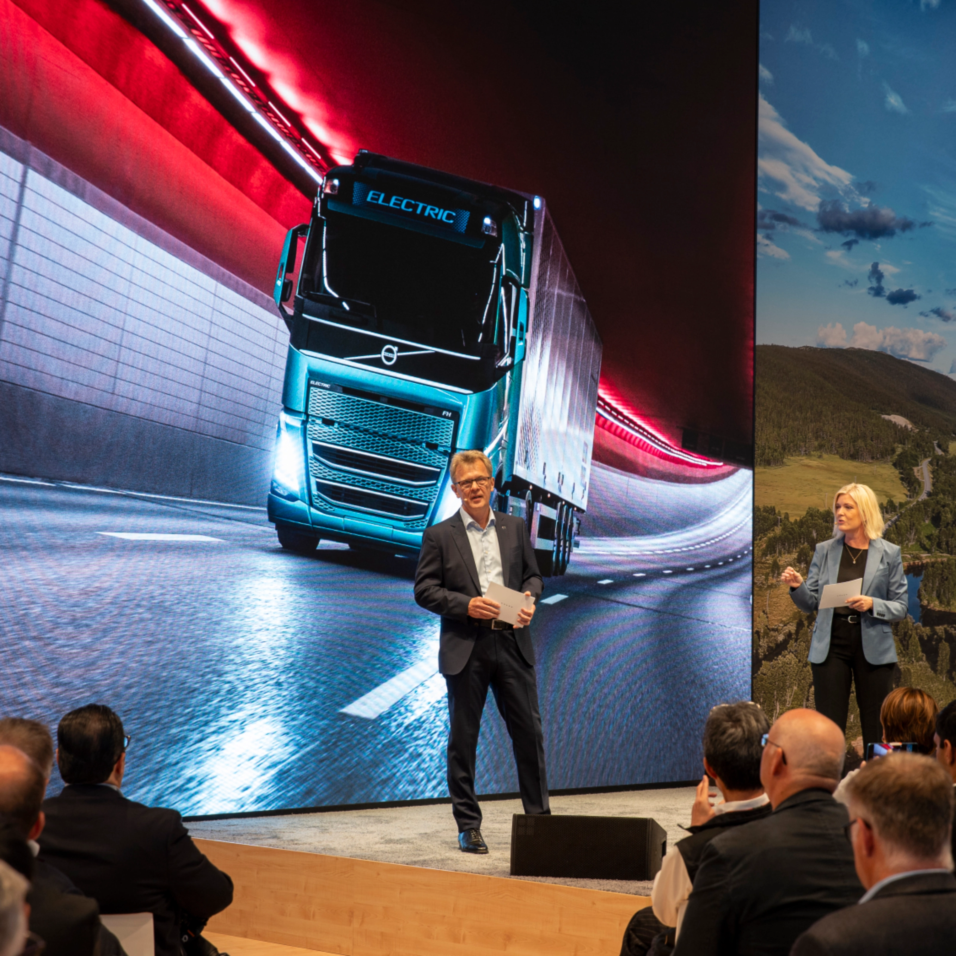 Zwei Redner präsentieren vor einer großen Leinwand mit einem E-Lkw in einem Tunnel.