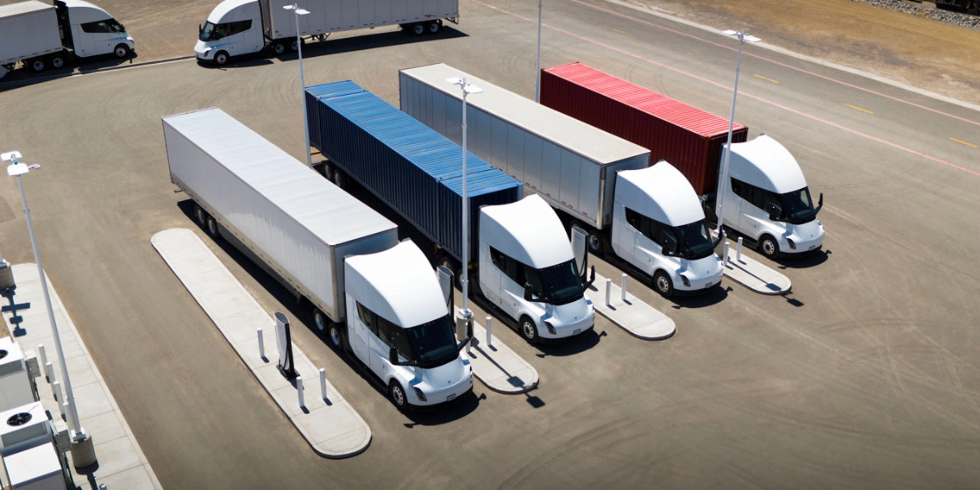 Several white electric semi-trucks are charging at a large station with colored containers.