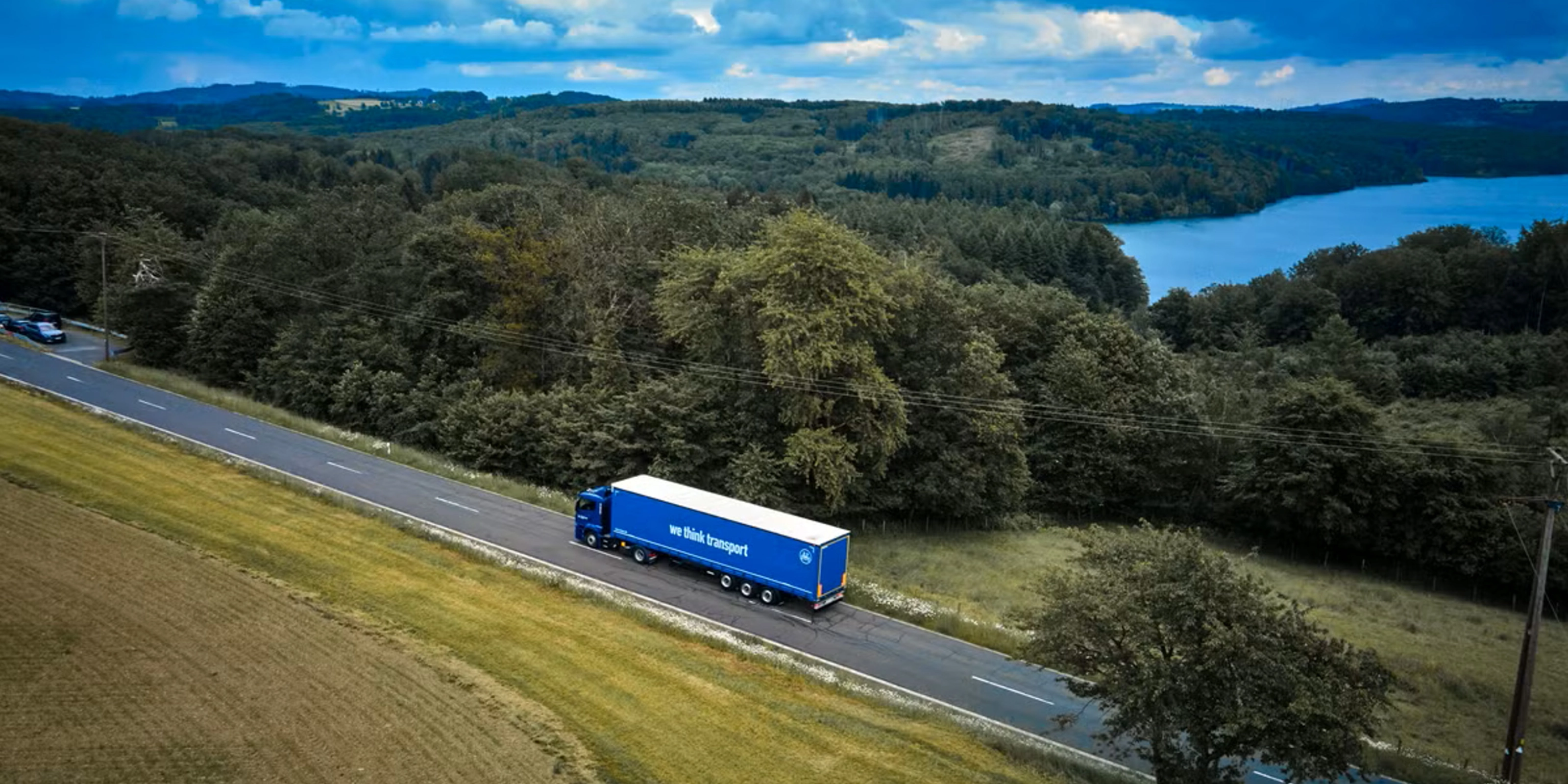 Blue BPW truck driving on a rural road beside a forest and lake.