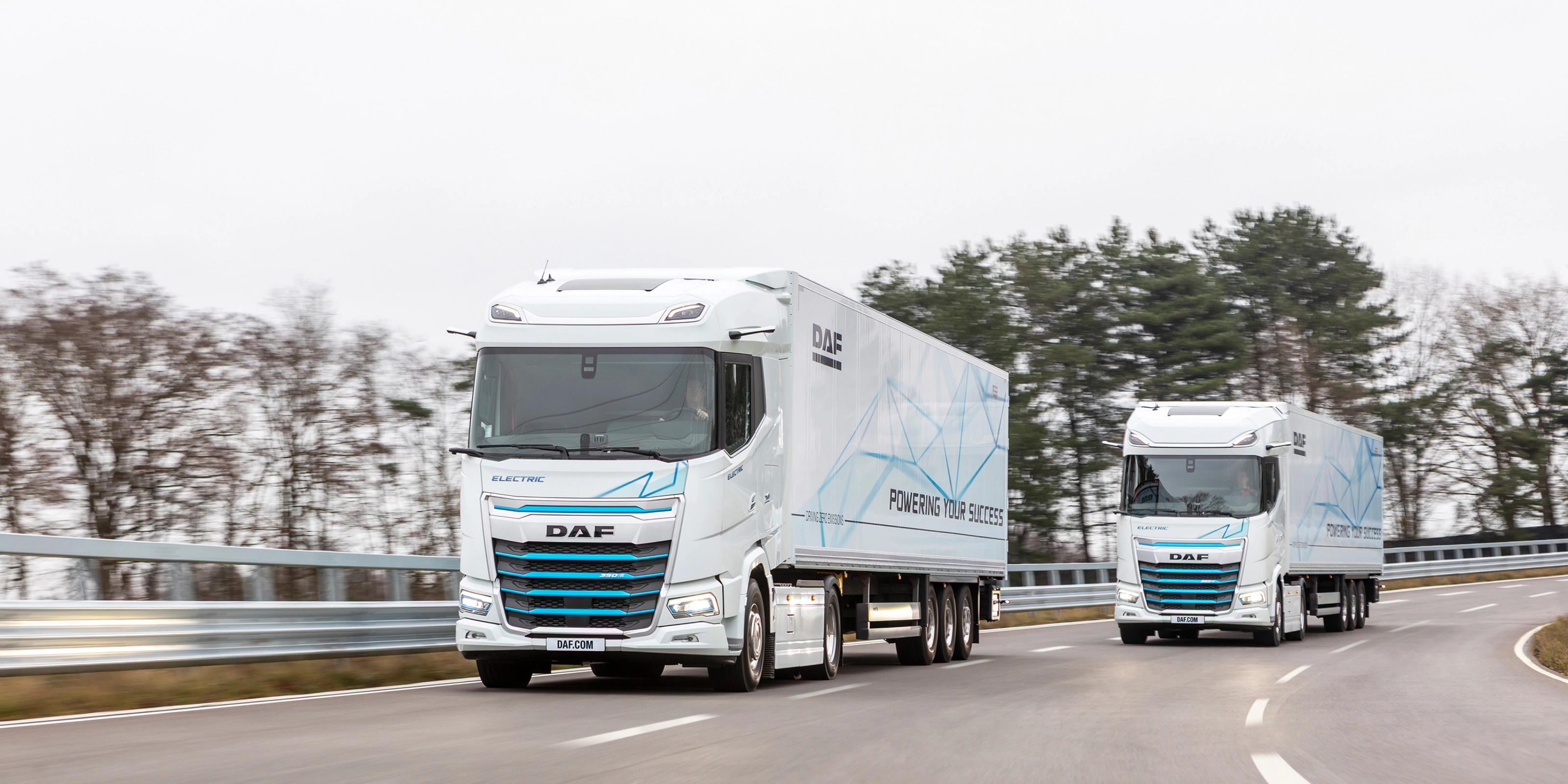 Two electric DAF trucks driving on a curved road with trees in the background.