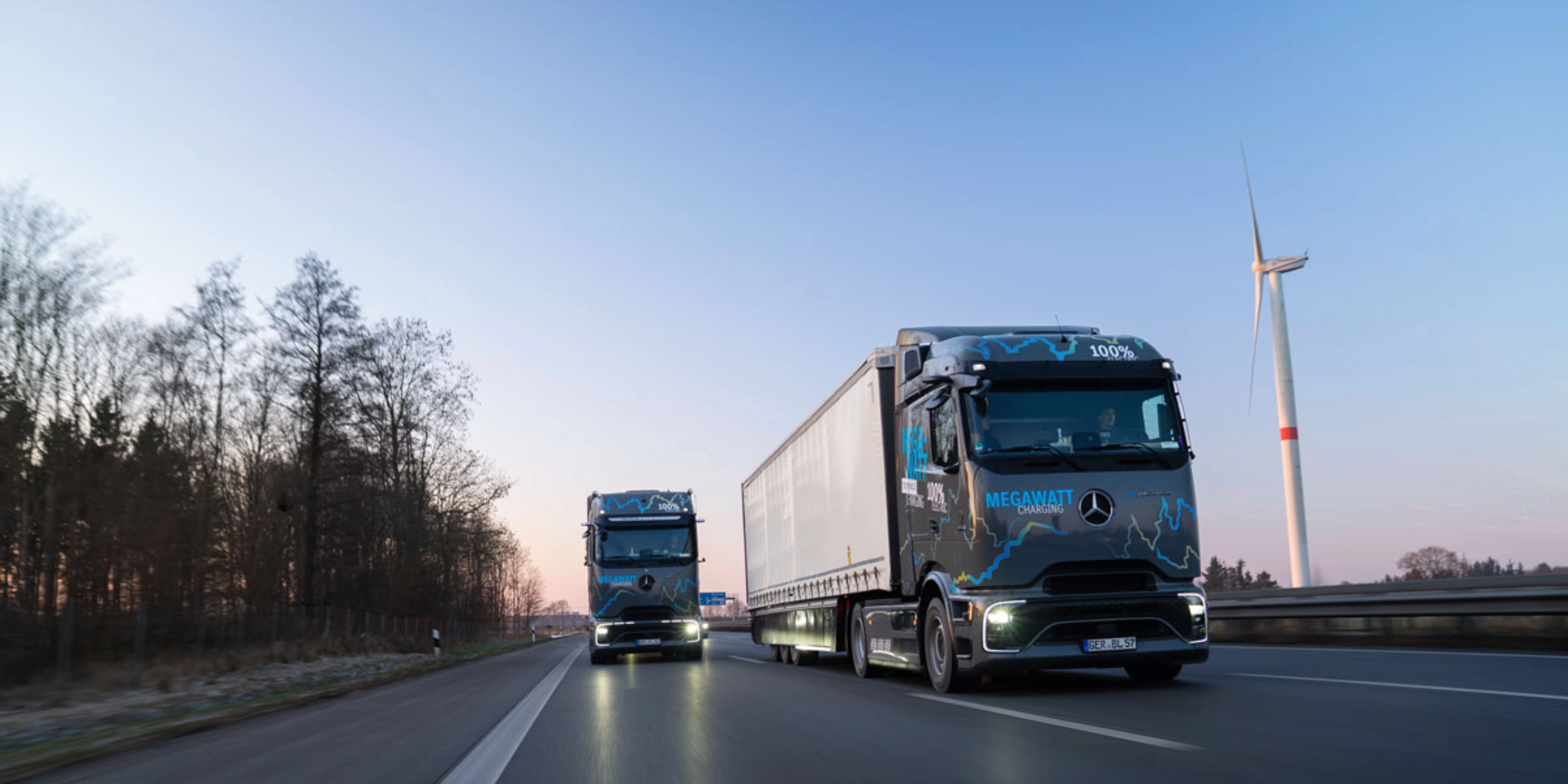 Zwei elektrische Mercedes-Benz-Lkw mit „Megawatt Charging“-Aufschrift fahren bei Dämmerung auf der Autobahn.