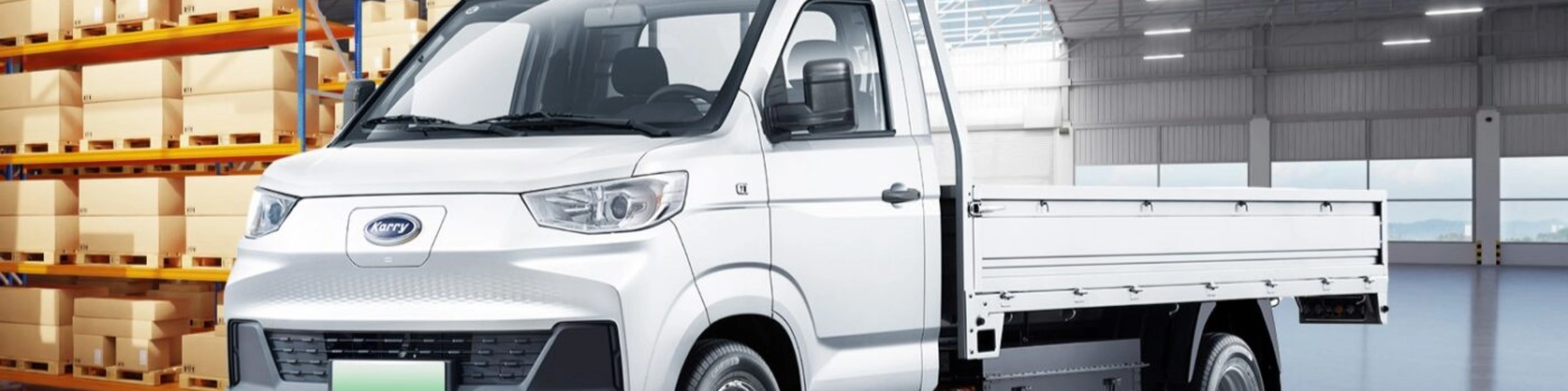 A white Karry electric flatbed truck is parked inside a warehouse near stacked boxes.