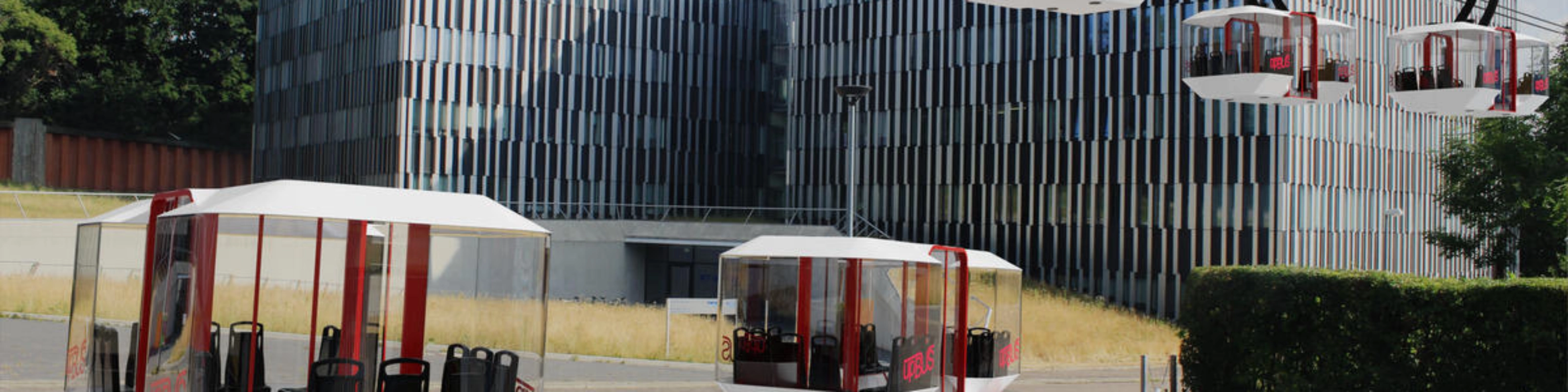 Moderne Seilbahnen und Bodenfahrzeuge des upBUS-Systems stehen vor Bürogebäuden.