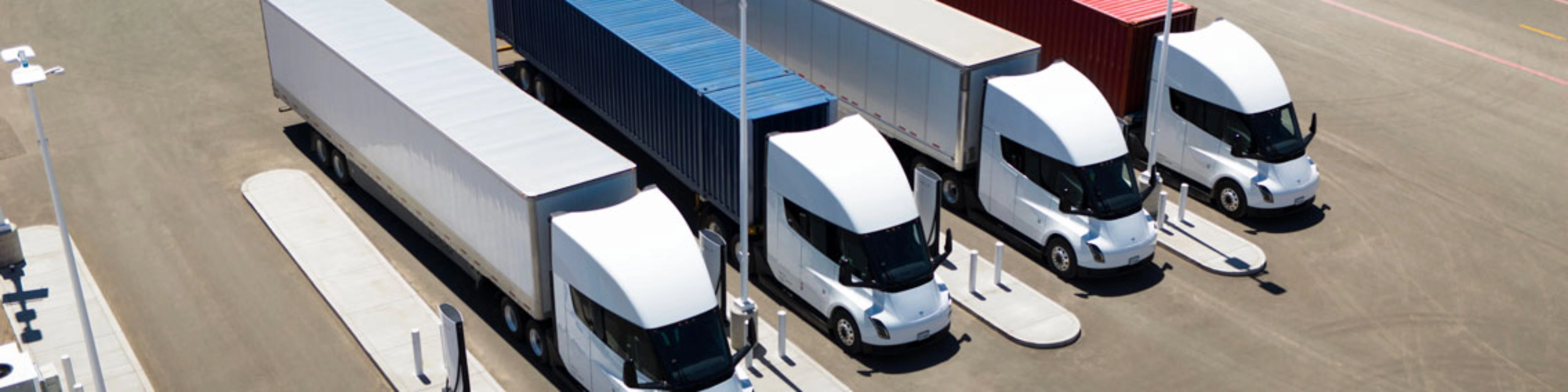 Several white electric semi-trucks are charging at a large station with colored containers.