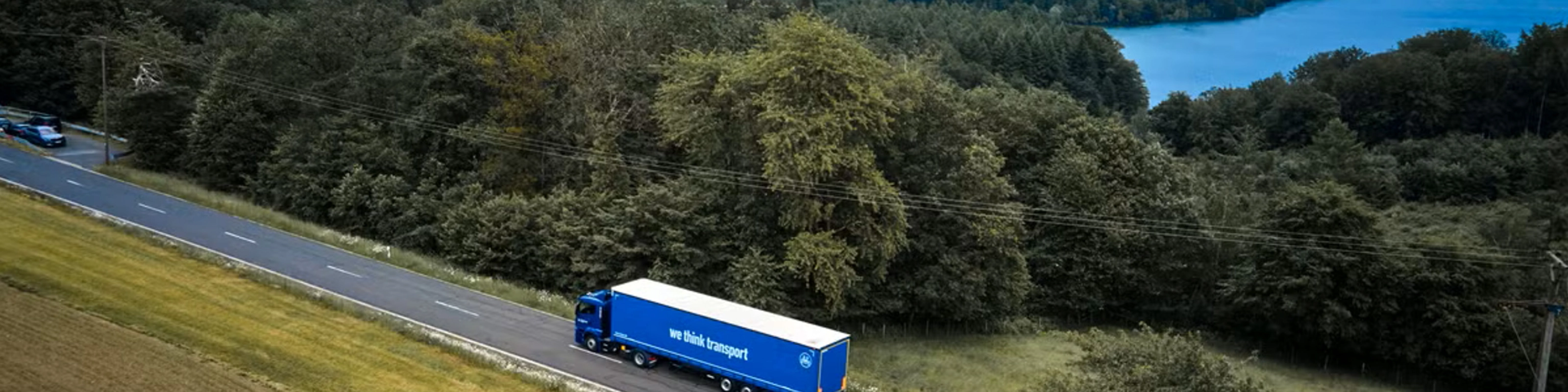 Blue BPW truck driving on a rural road beside a forest and lake.