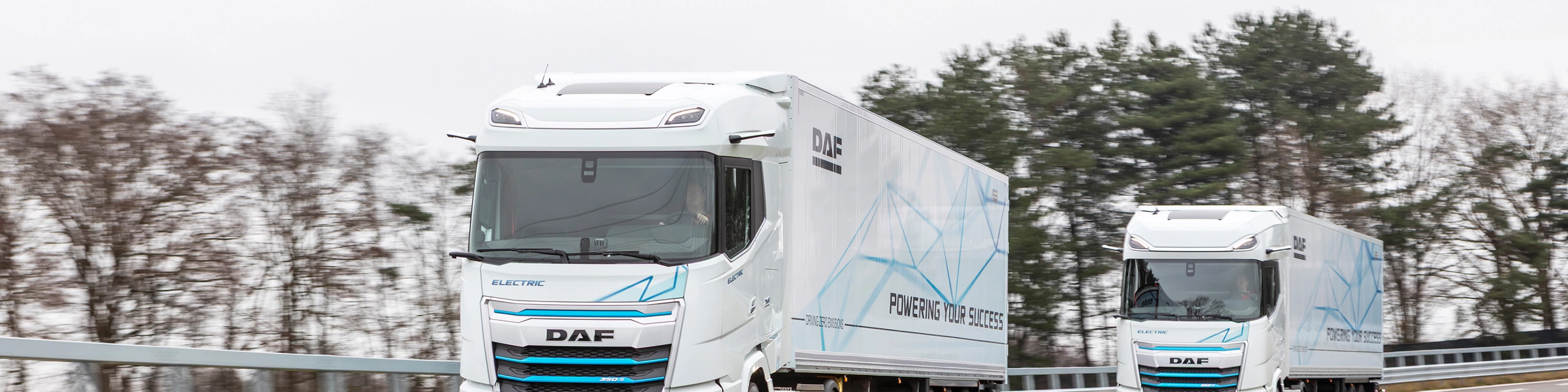 Two electric DAF trucks driving on a curved road with trees in the background.