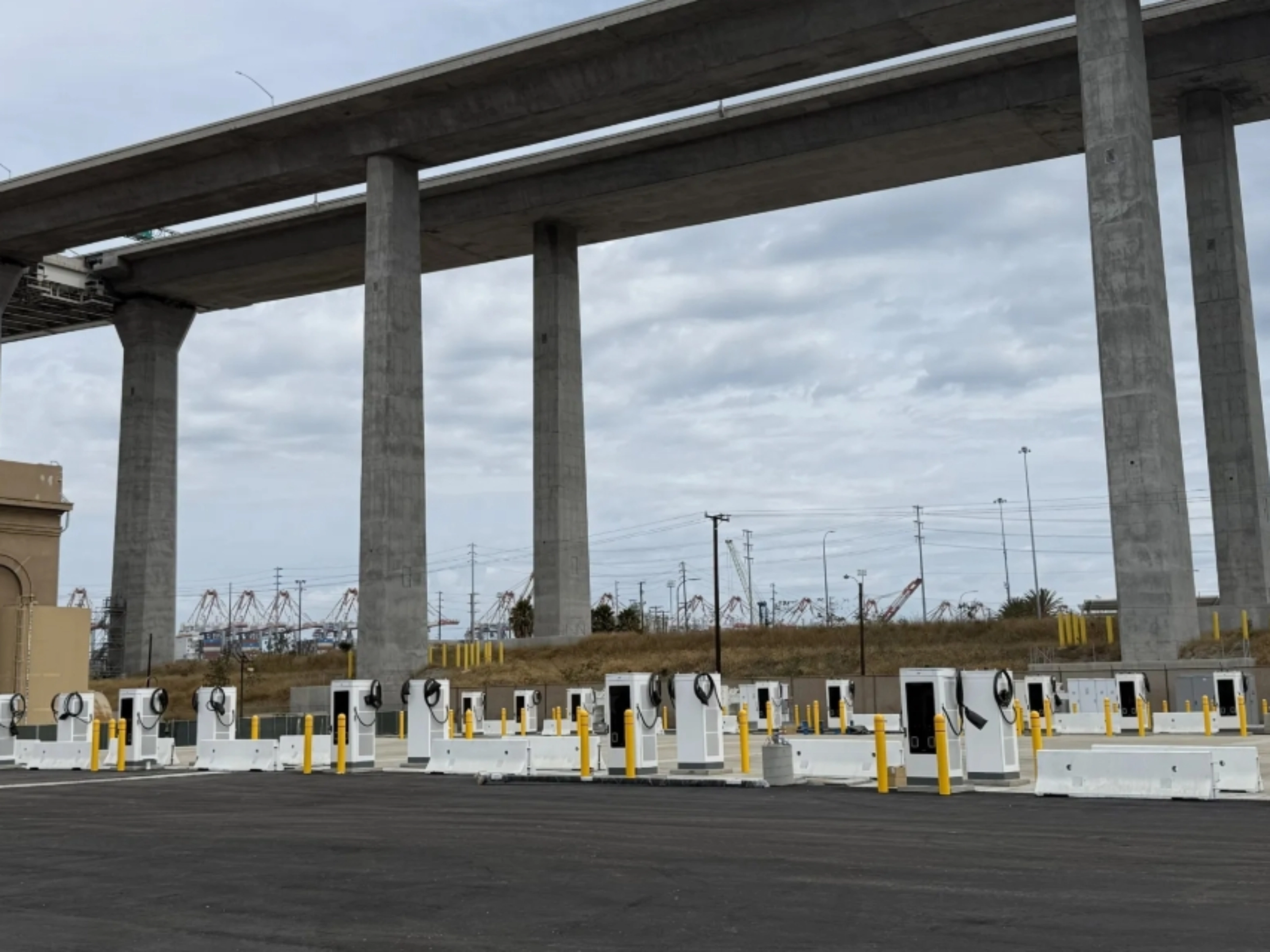 Mehrere E-Ladestationen auf einem Parkplatz unter einer Autobahnbrücke.