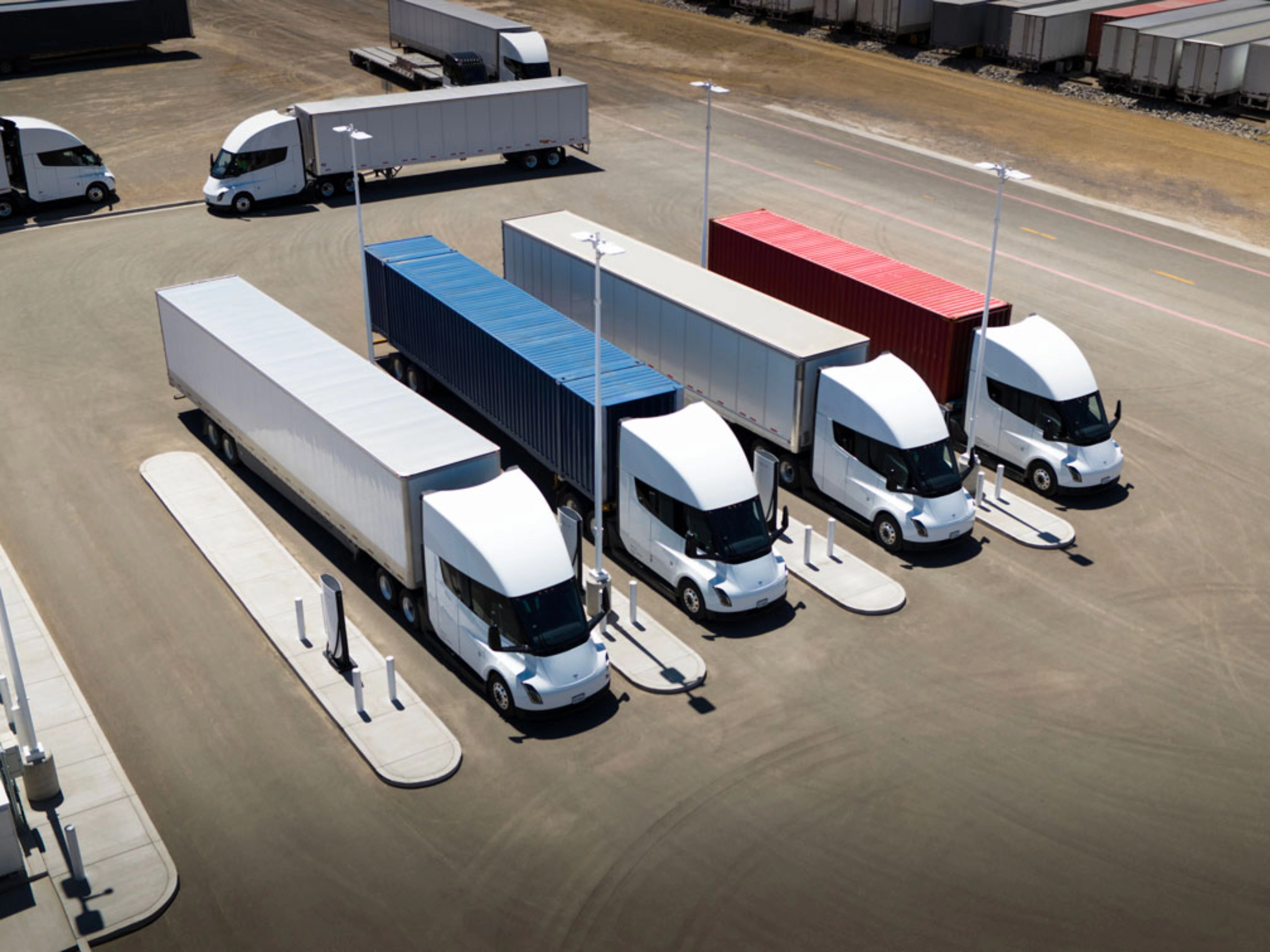 Several white electric semi-trucks are charging at a large station with colored containers.