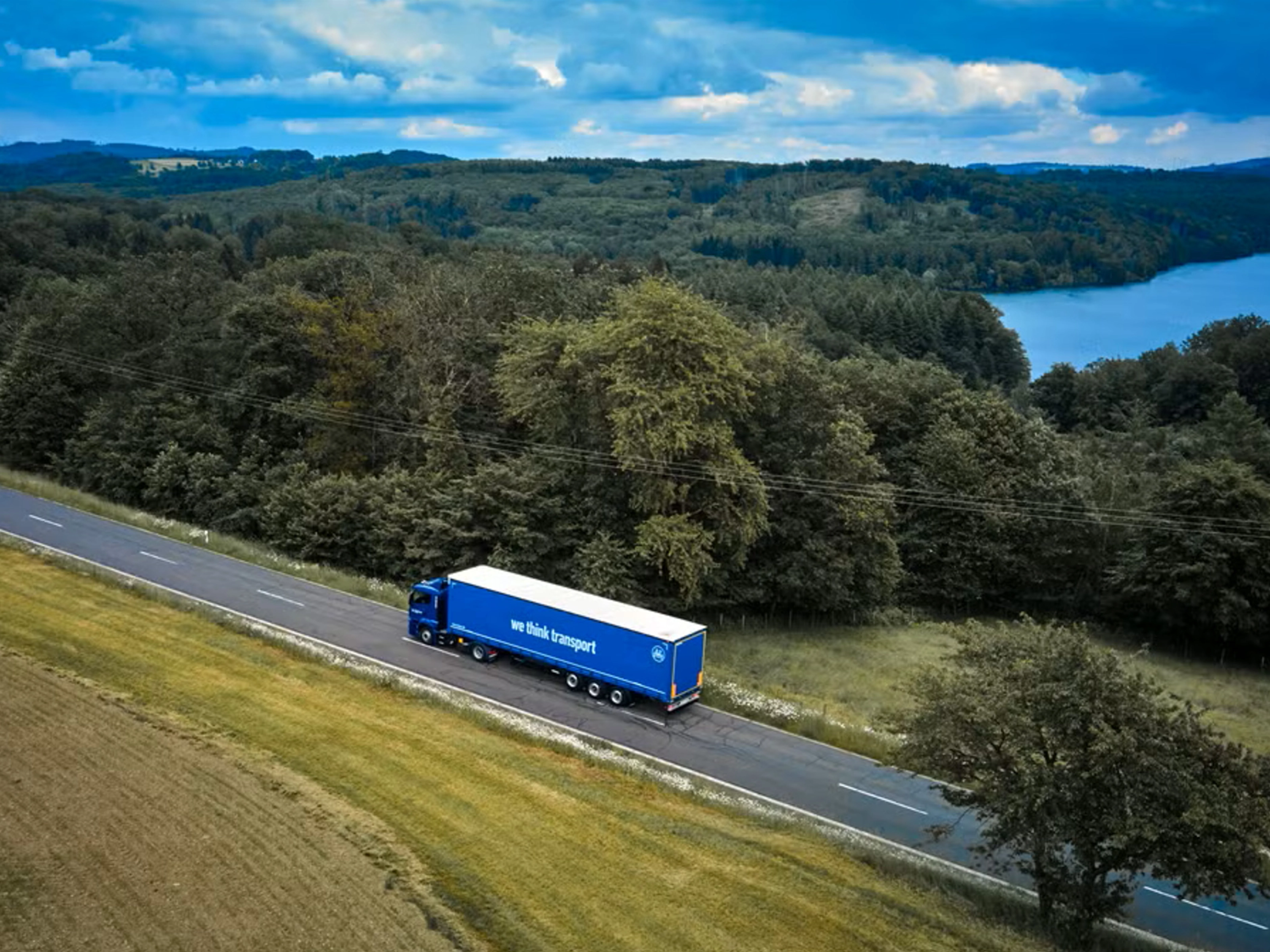 Blue BPW truck driving on a rural road beside a forest and lake.