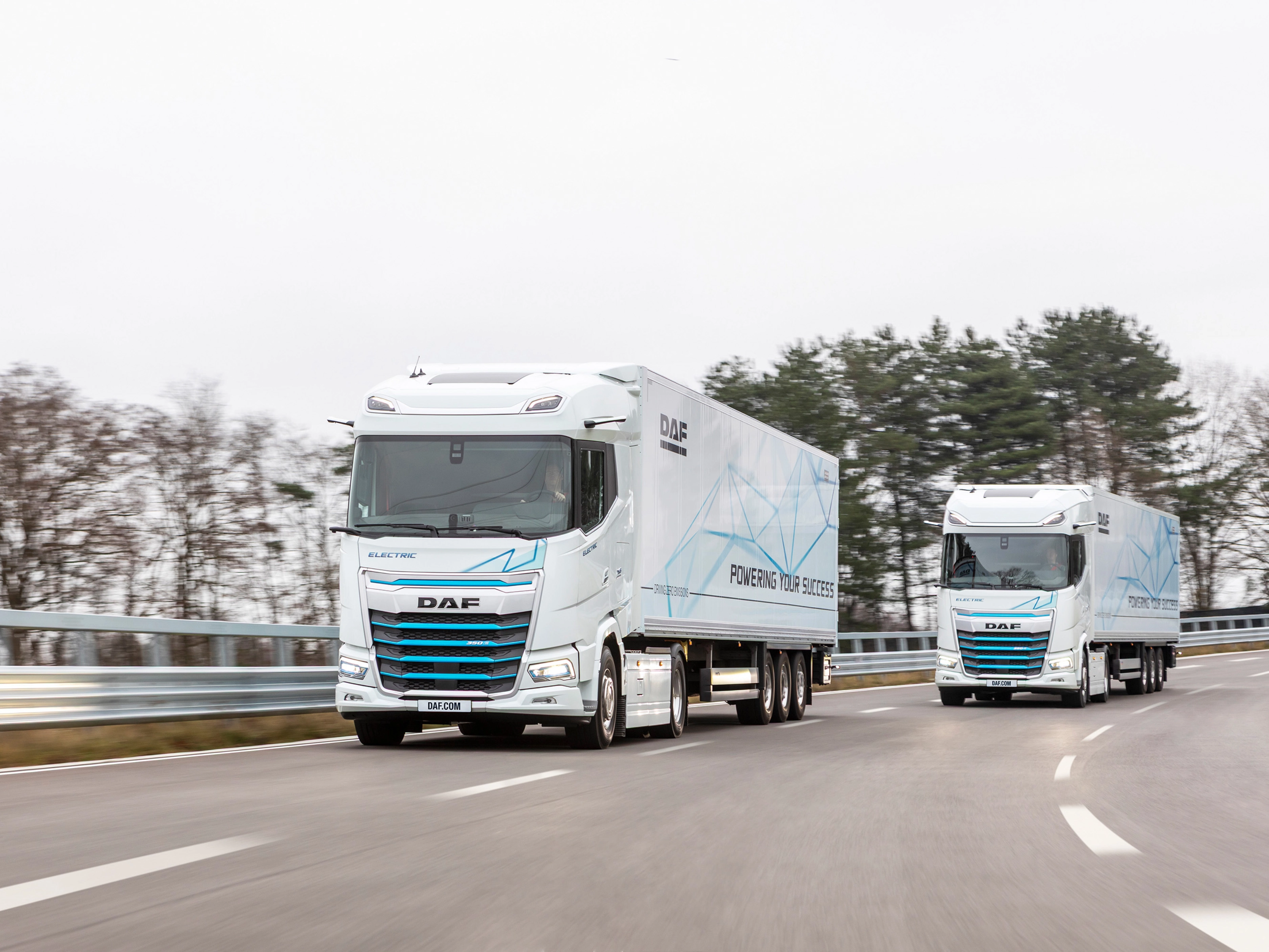 Two electric DAF trucks driving on a curved road with trees in the background.