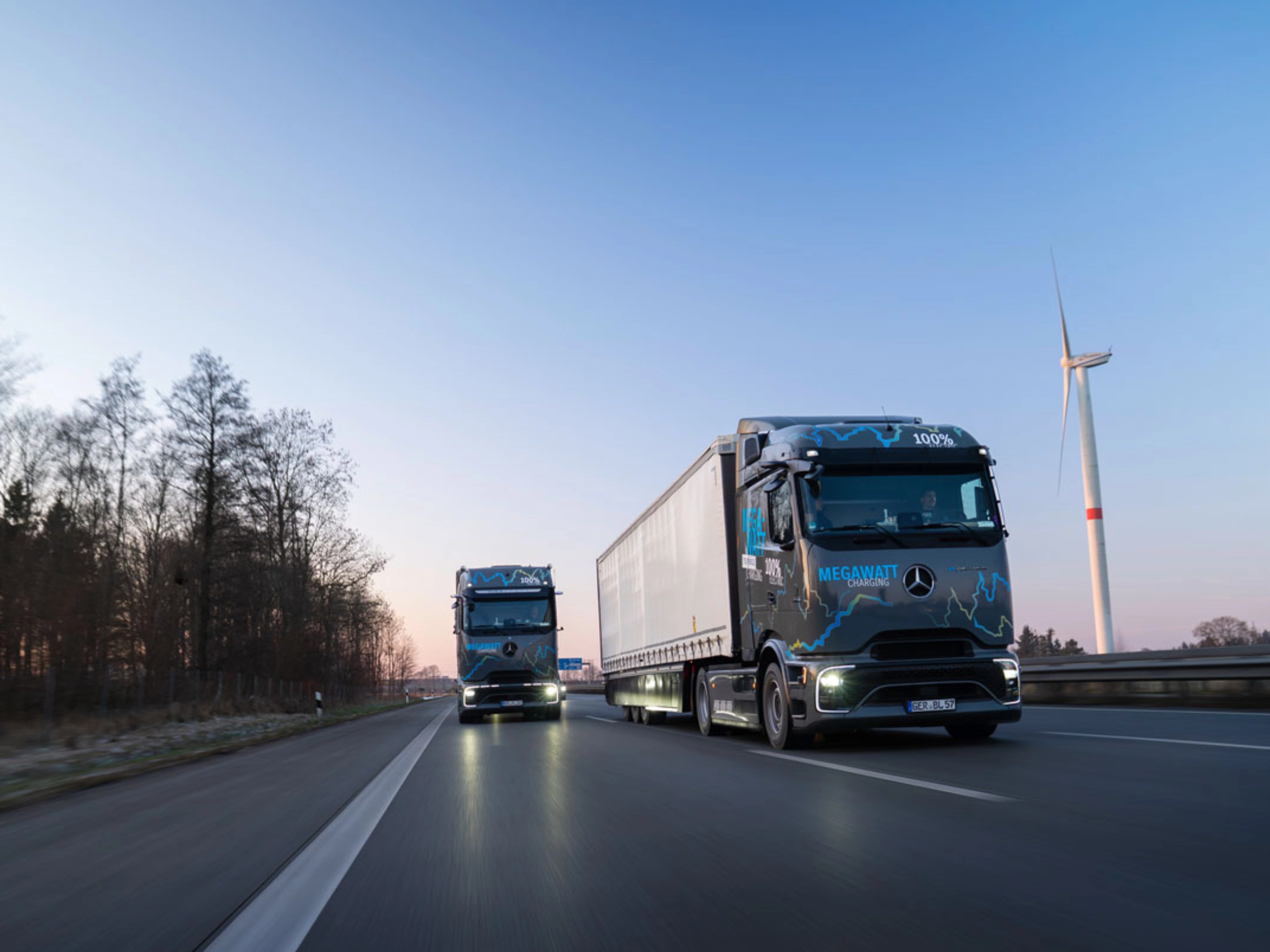 Zwei elektrische Mercedes-Benz-Lkw mit „Megawatt Charging“-Aufschrift fahren bei Dämmerung auf der Autobahn.