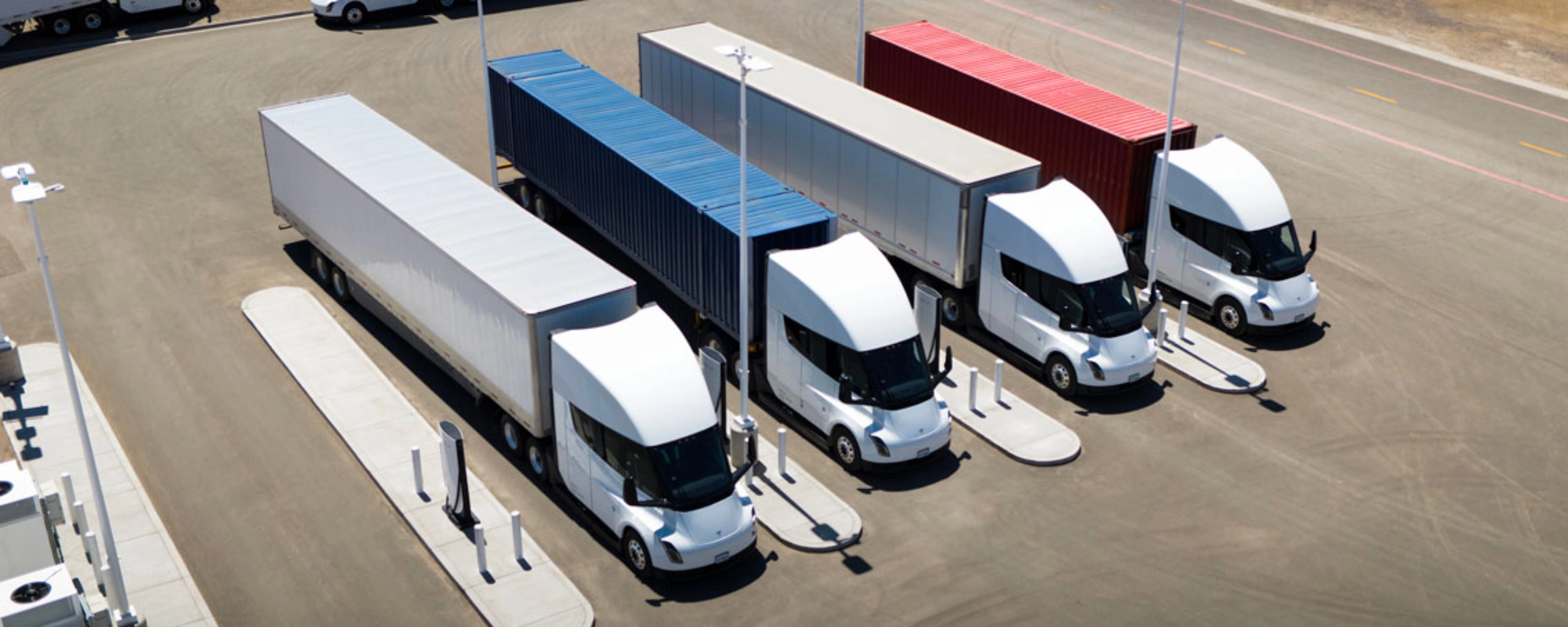 Several white electric semi-trucks are charging at a large station with colored containers.