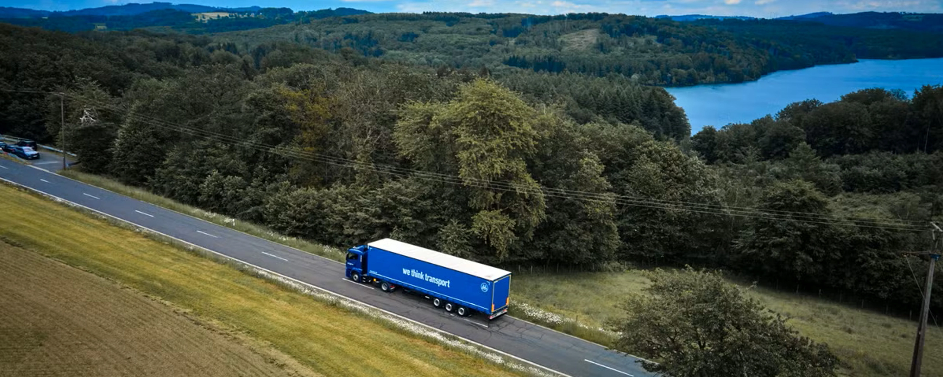 Blue BPW truck driving on a rural road beside a forest and lake.