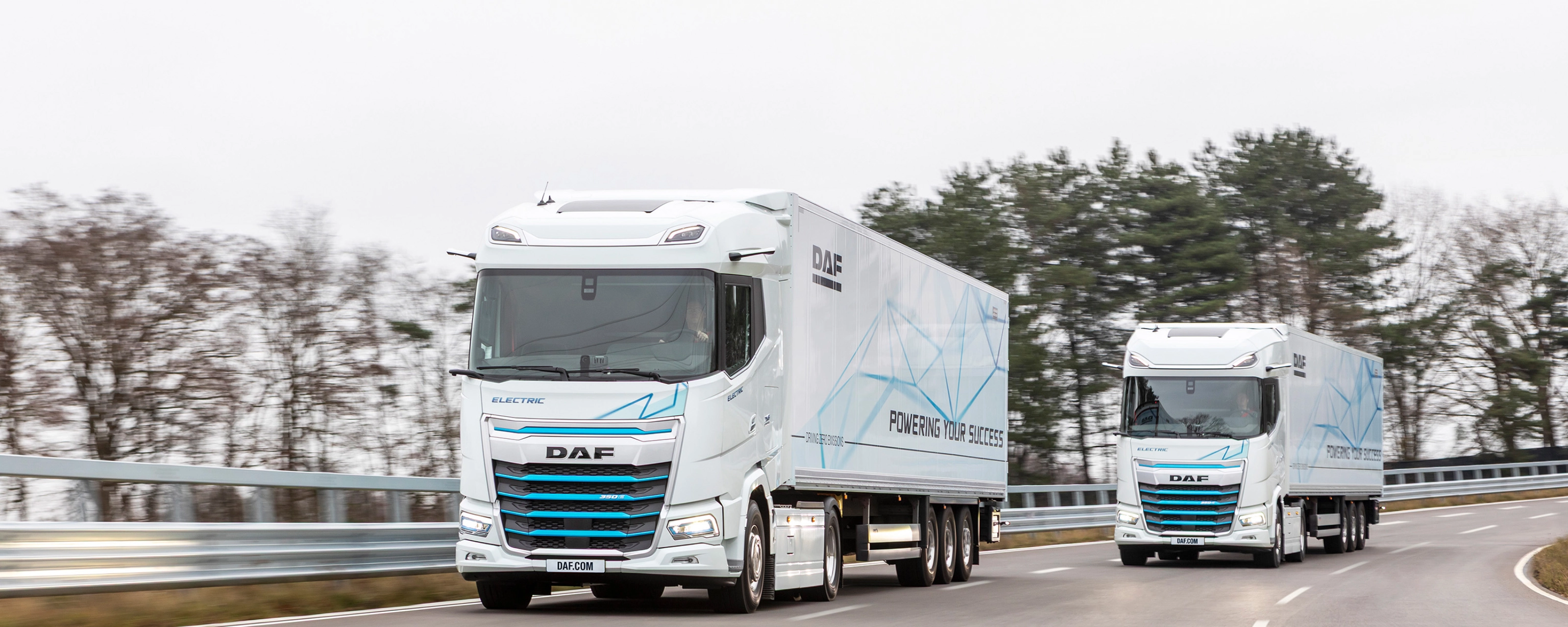 Two electric DAF trucks driving on a curved road with trees in the background.