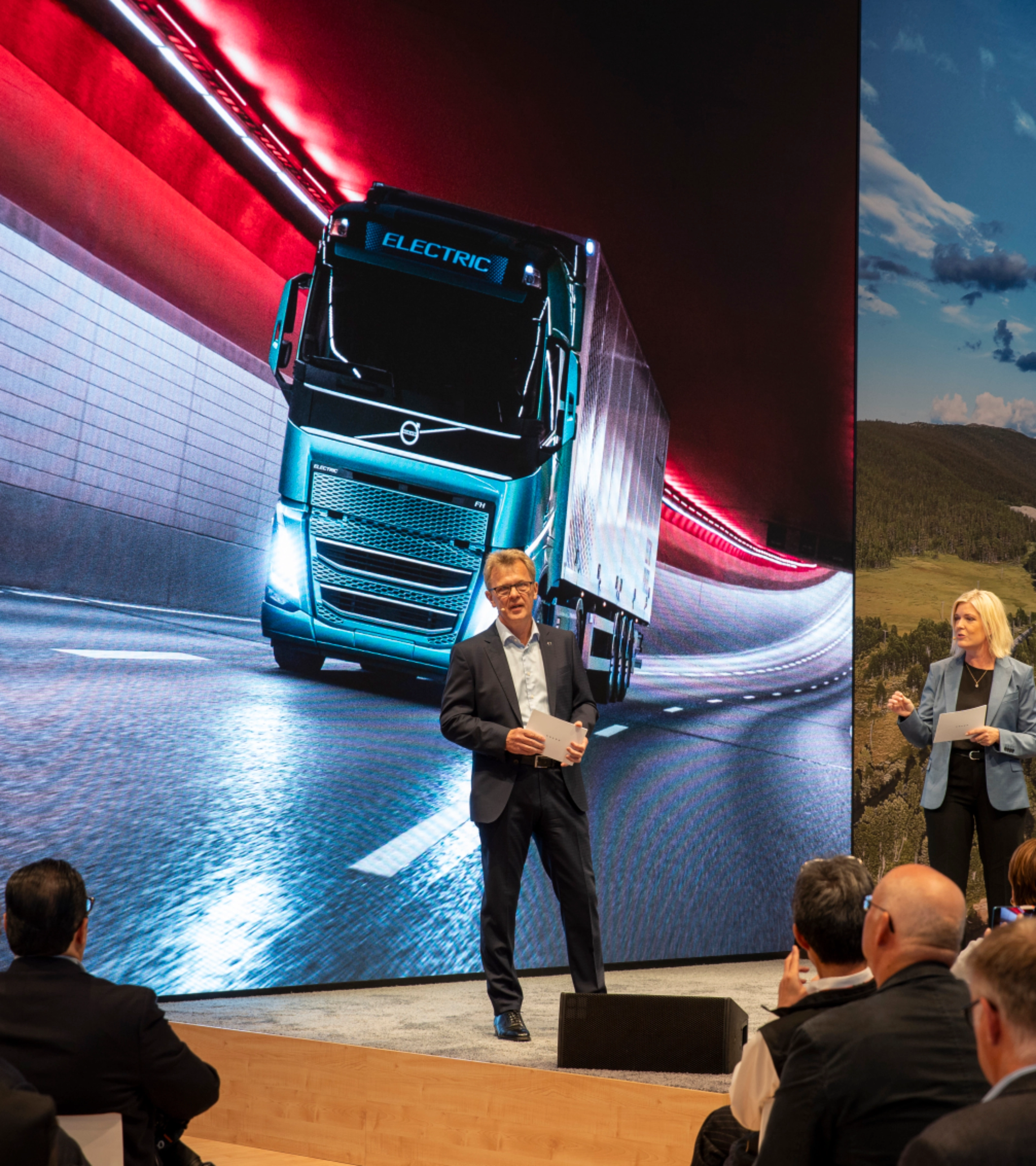 Zwei Redner präsentieren vor einer großen Leinwand mit einem E-Lkw in einem Tunnel.