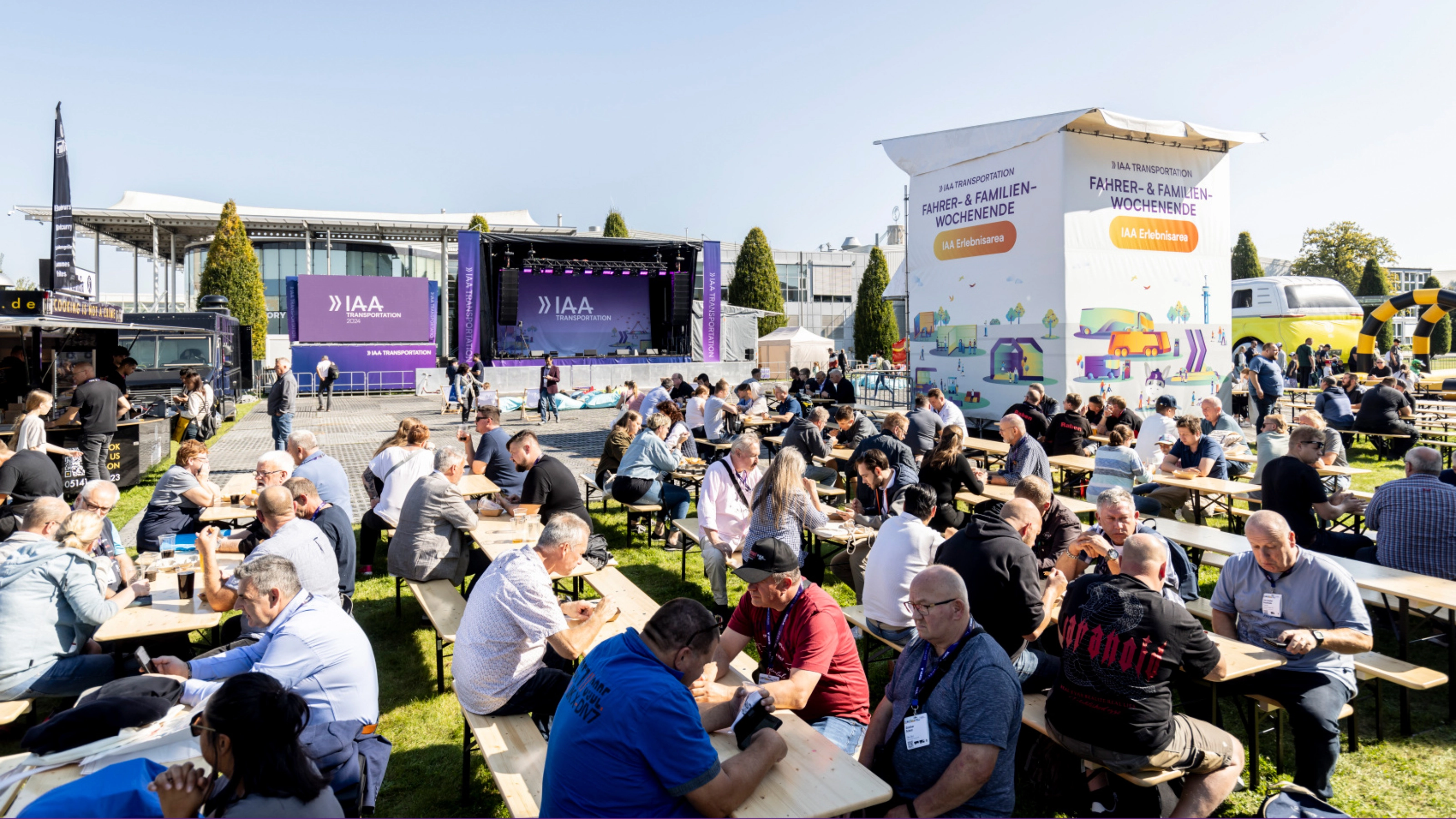 Besucher sitzen an Biertischen vor einer Bühne bei einer Freiluftveranstaltung.
