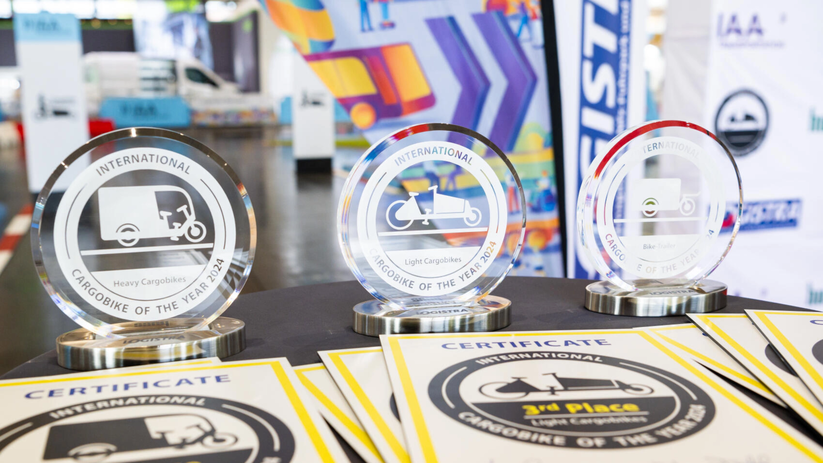 Drei Trophäen und Urkunden der Auszeichnung „International Cargobike of the Year 2024“ auf einem Tisch.
