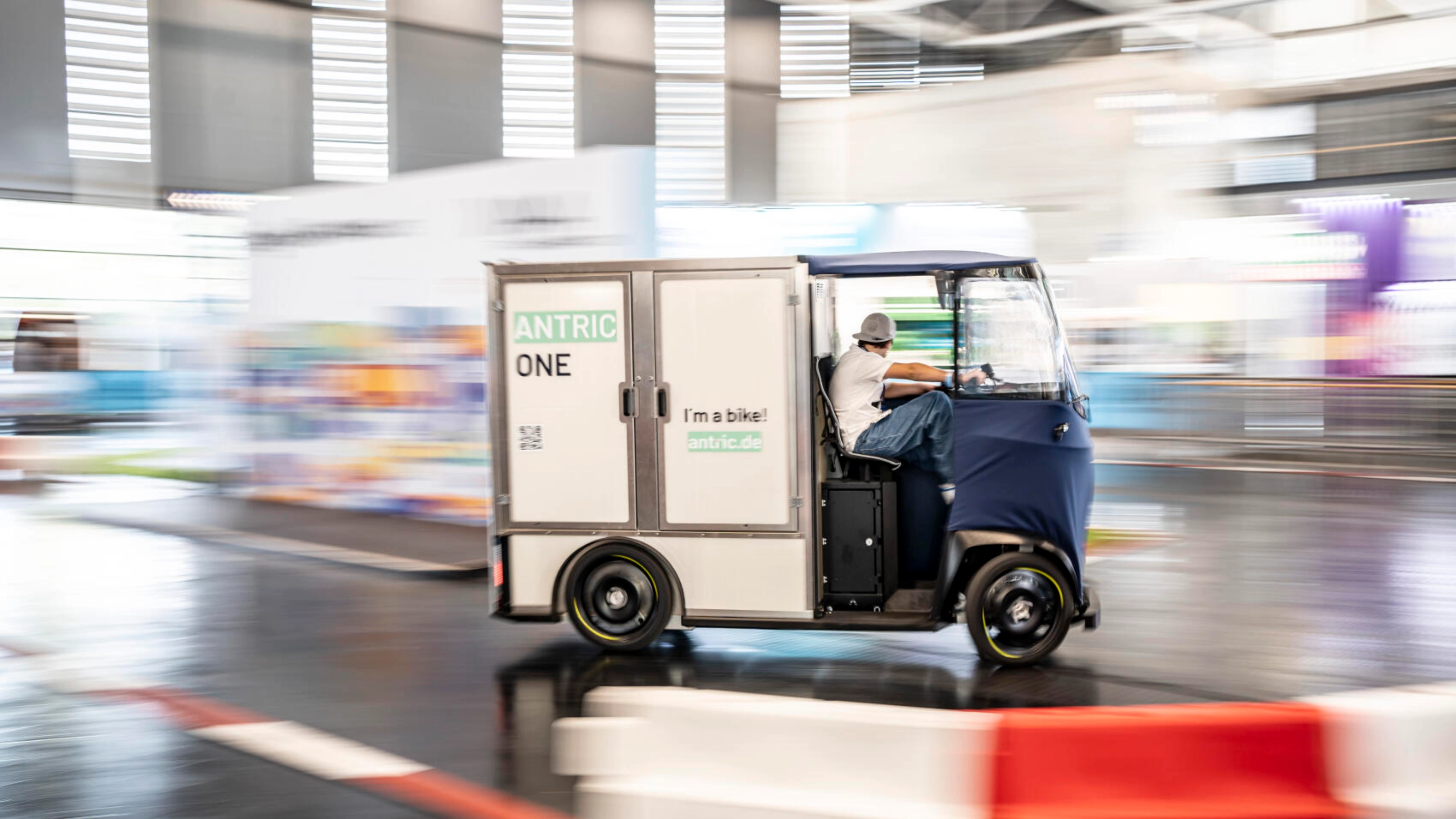 Kleines Lastenradfahrzeug mit Aufschrift ANTRIC ONE fährt in einer Messehalle.
