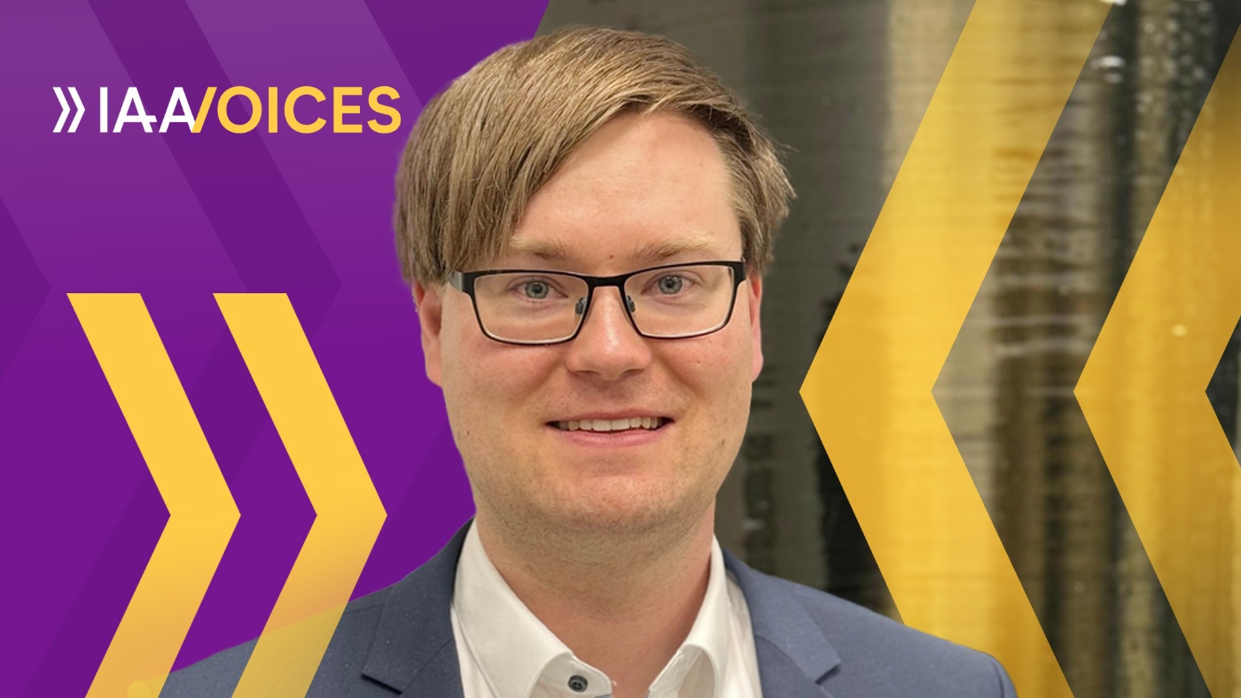 Porträt von Patrick Seidel lächelnd mit Brille vor IAA-Voices-Hintergrund.