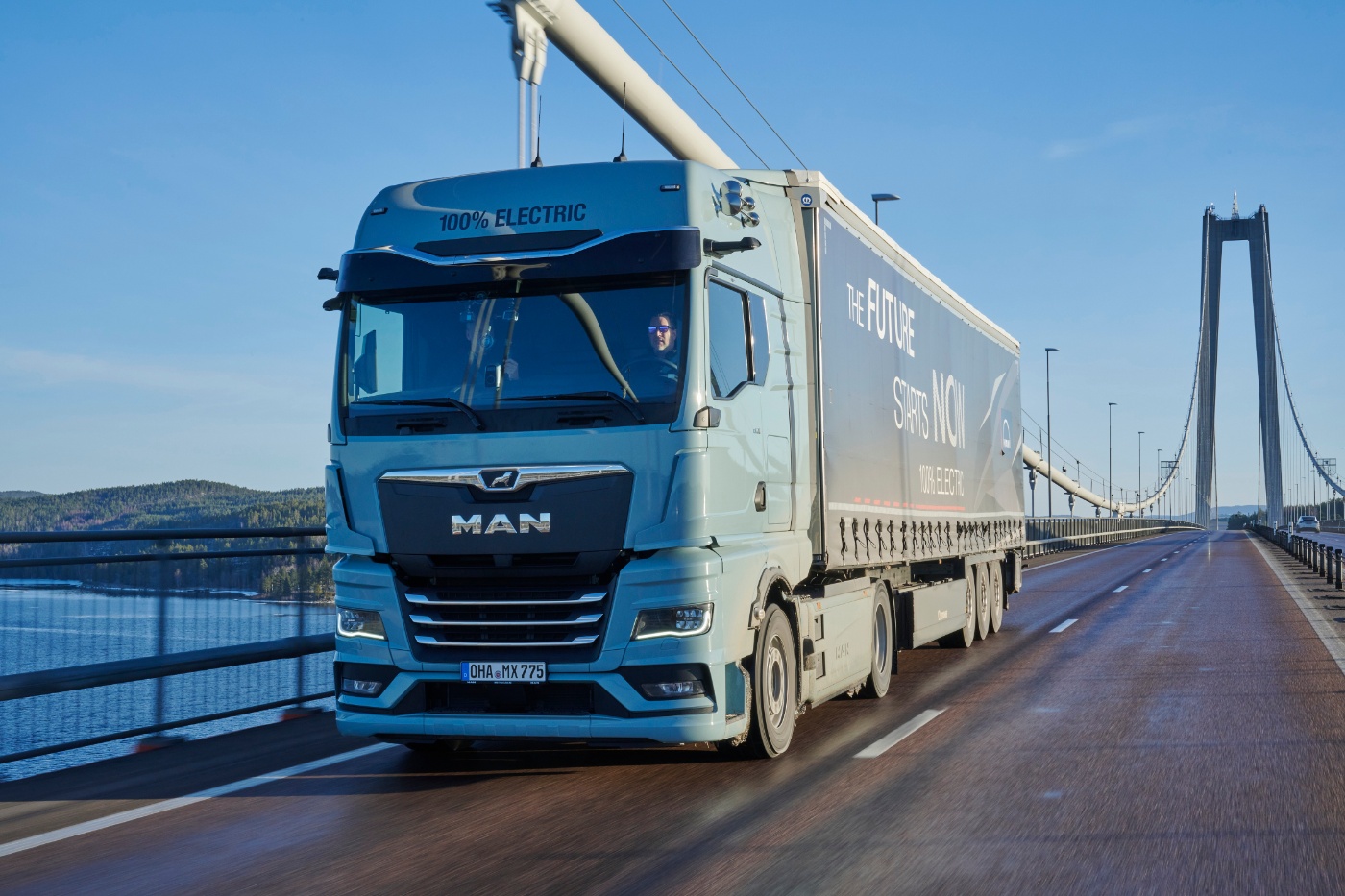 Hellblauer MAN Elektro-Lkw mit einem Anhänger mit der Aufschrift „The Future Starts Now“ fährt über eine Hängebrücke, im Hintergrund Wasser und Wald.